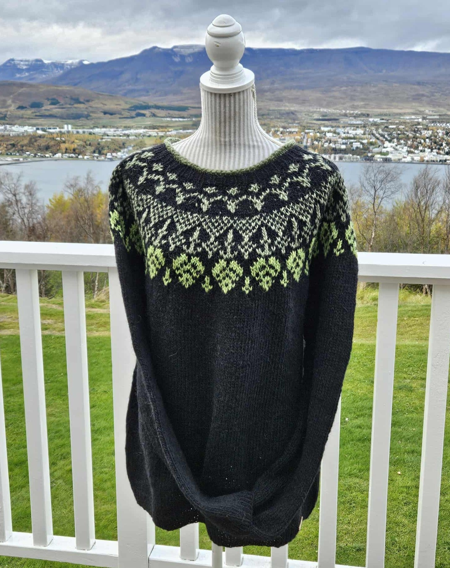 Black sweater with green patterns on a mannequin against a scenic backdrop of mountains and water.