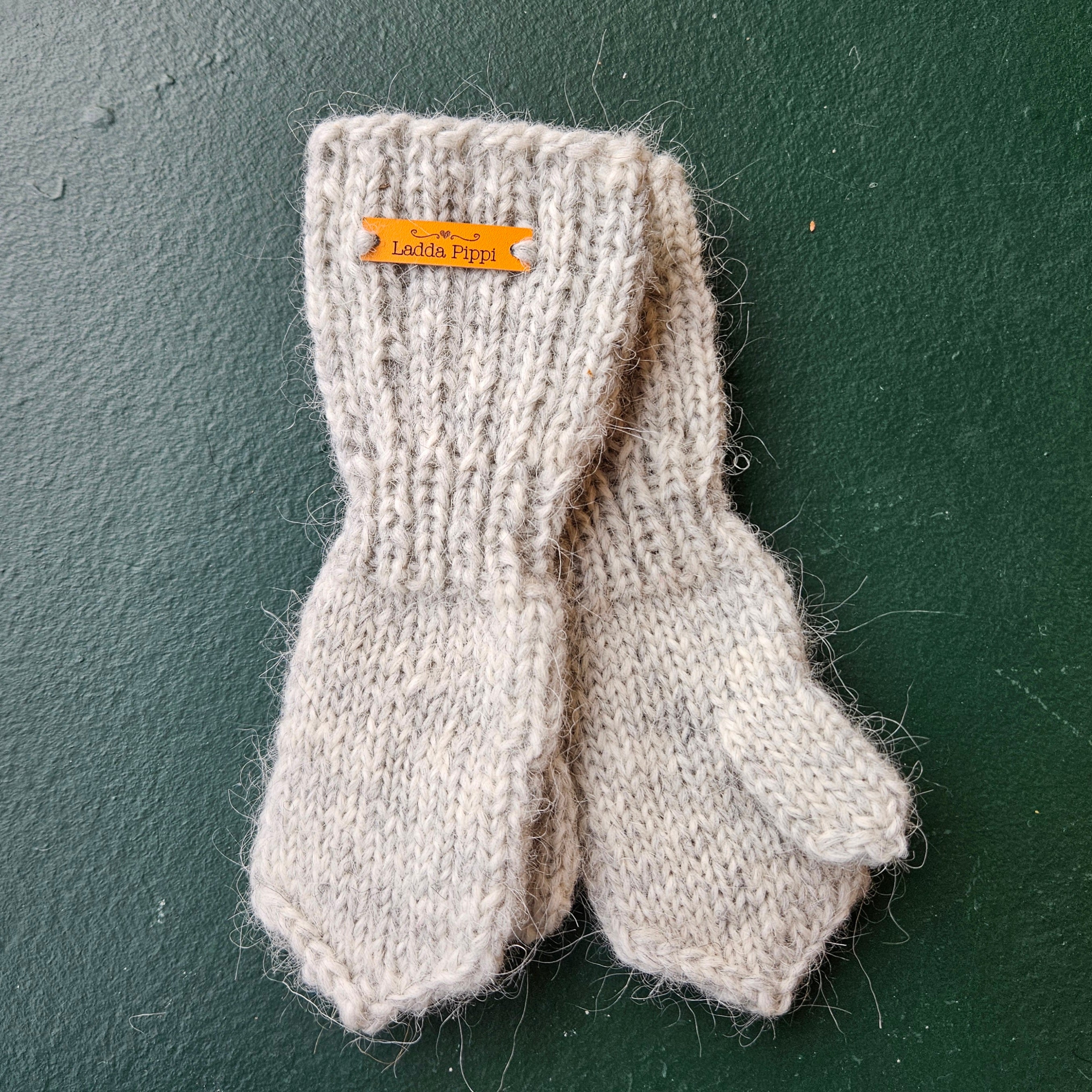 Pair of beige knitted mittens with a brand tag on a dark green background