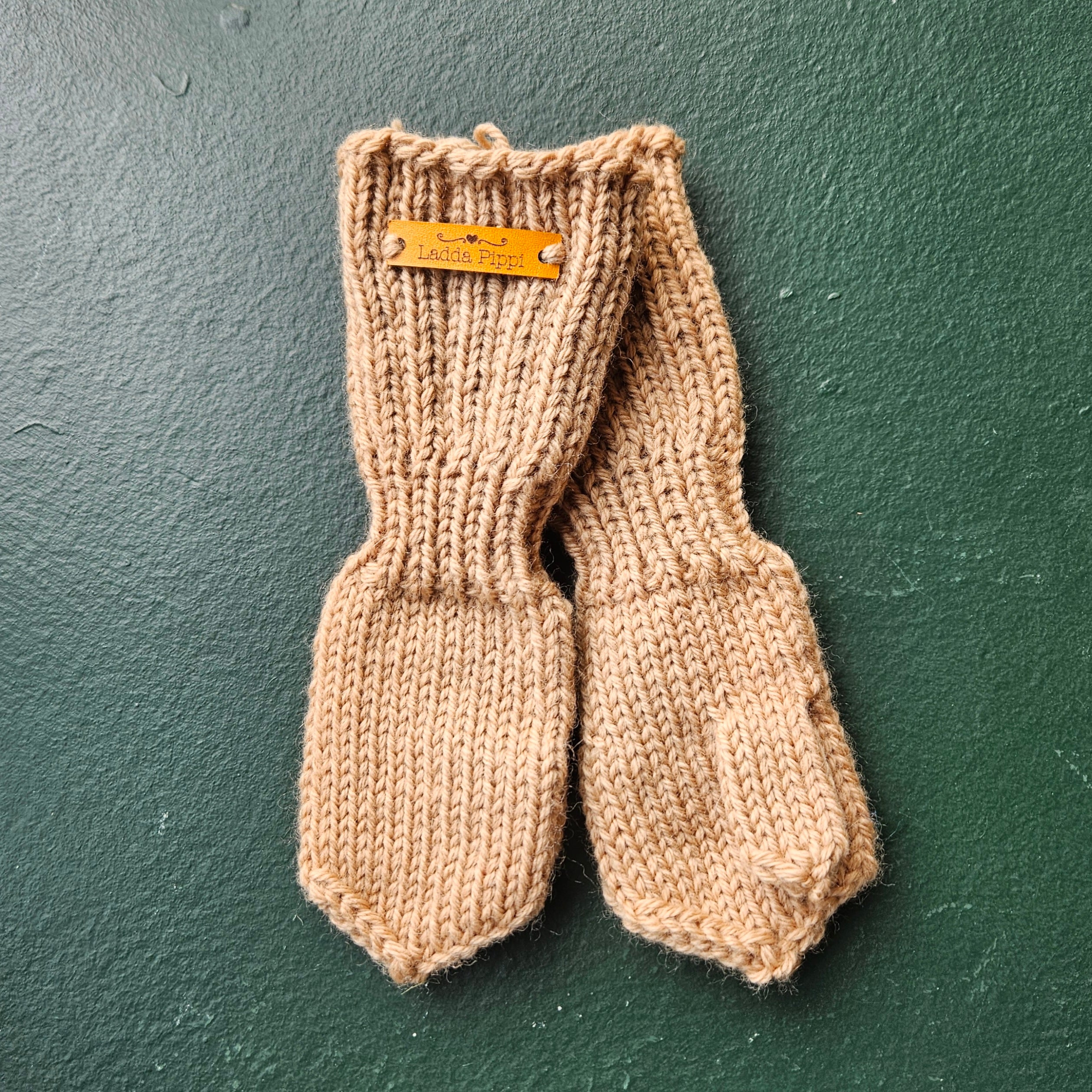 Pair of beige knitted baby mittens with a brand tag on a green background