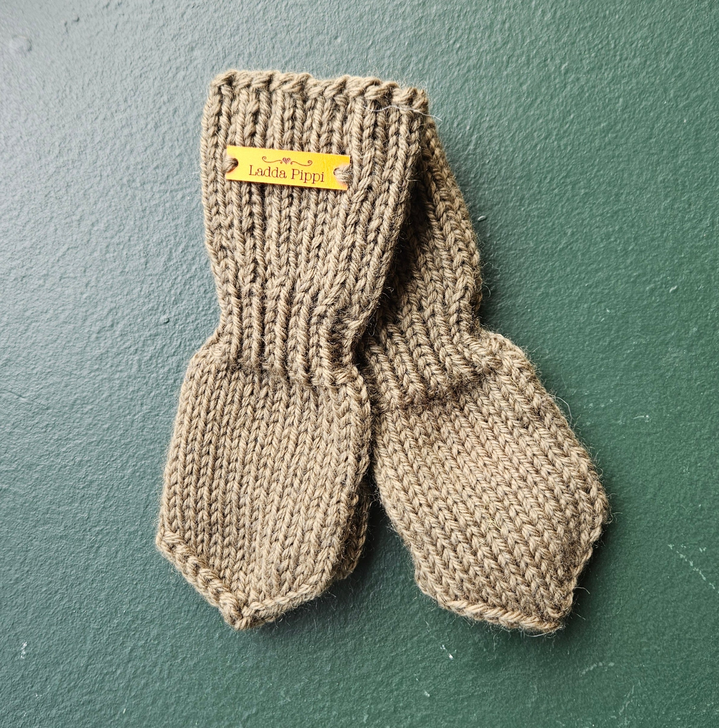 Pair of olive knitted baby mittens with a label on a green background