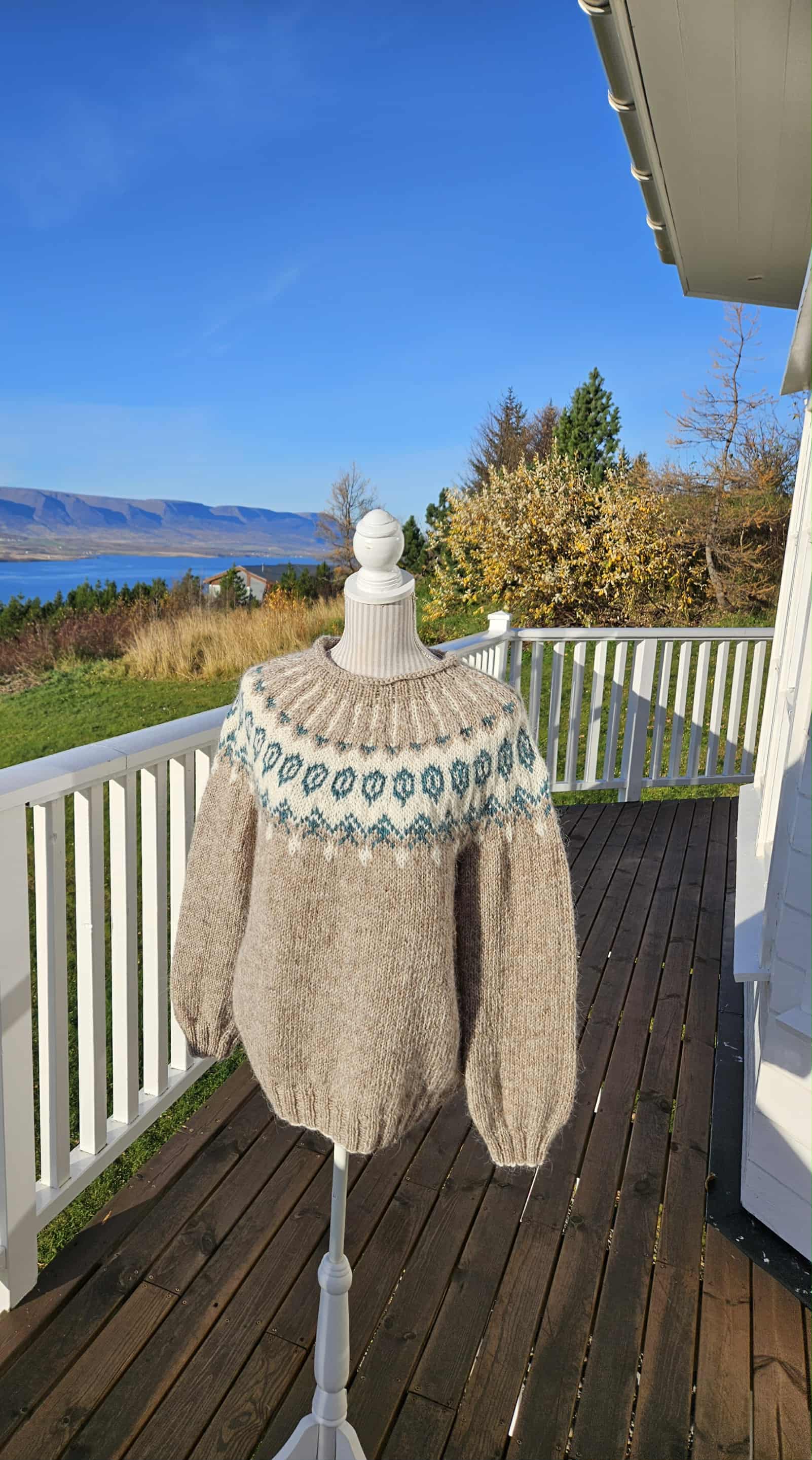 Beige sweater with a pattern on a mannequin stand on a wooden deck with a scenic background.