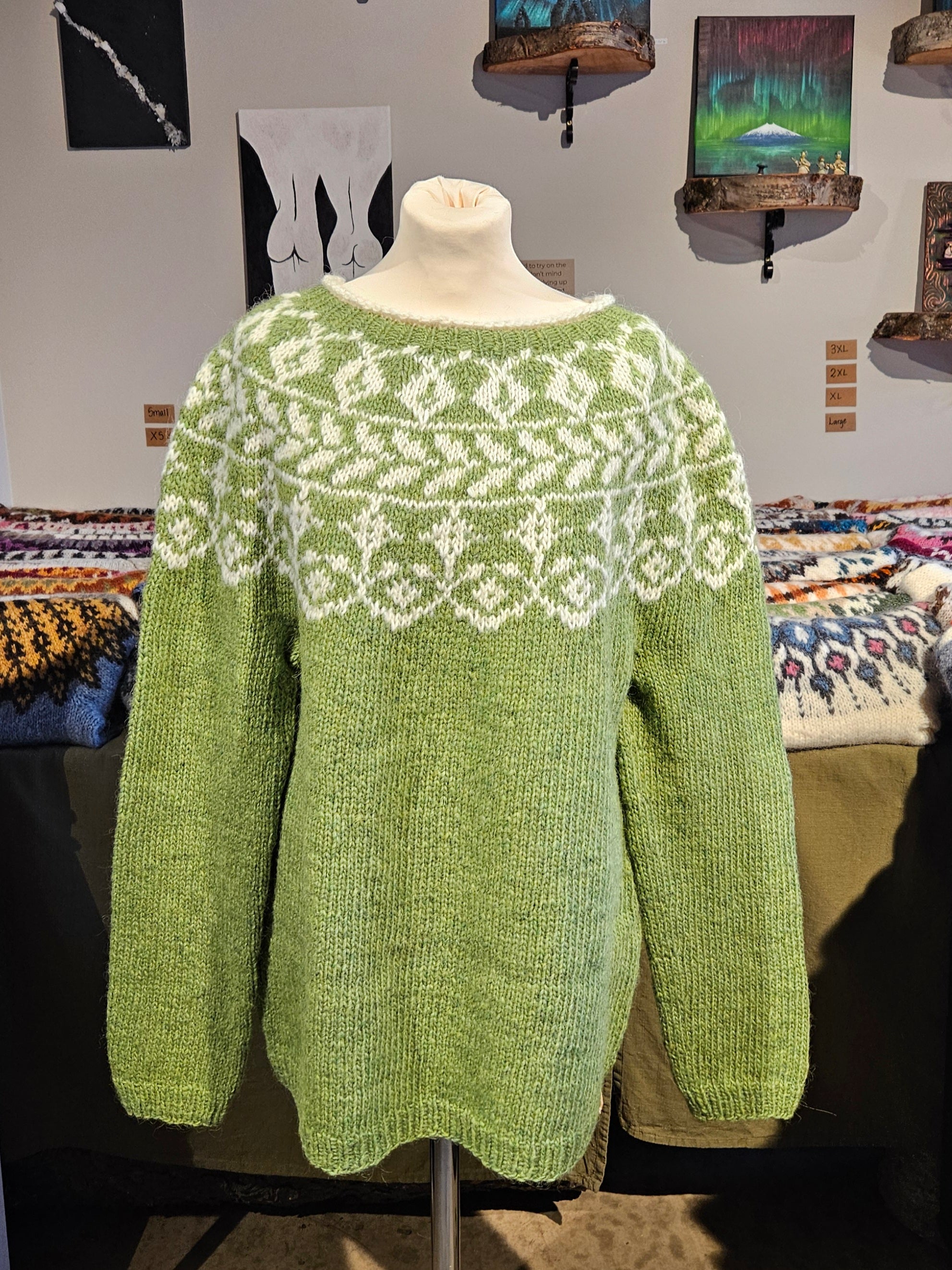 Green knitted sweater with white pattern on a mannequin against a wall with paintings.