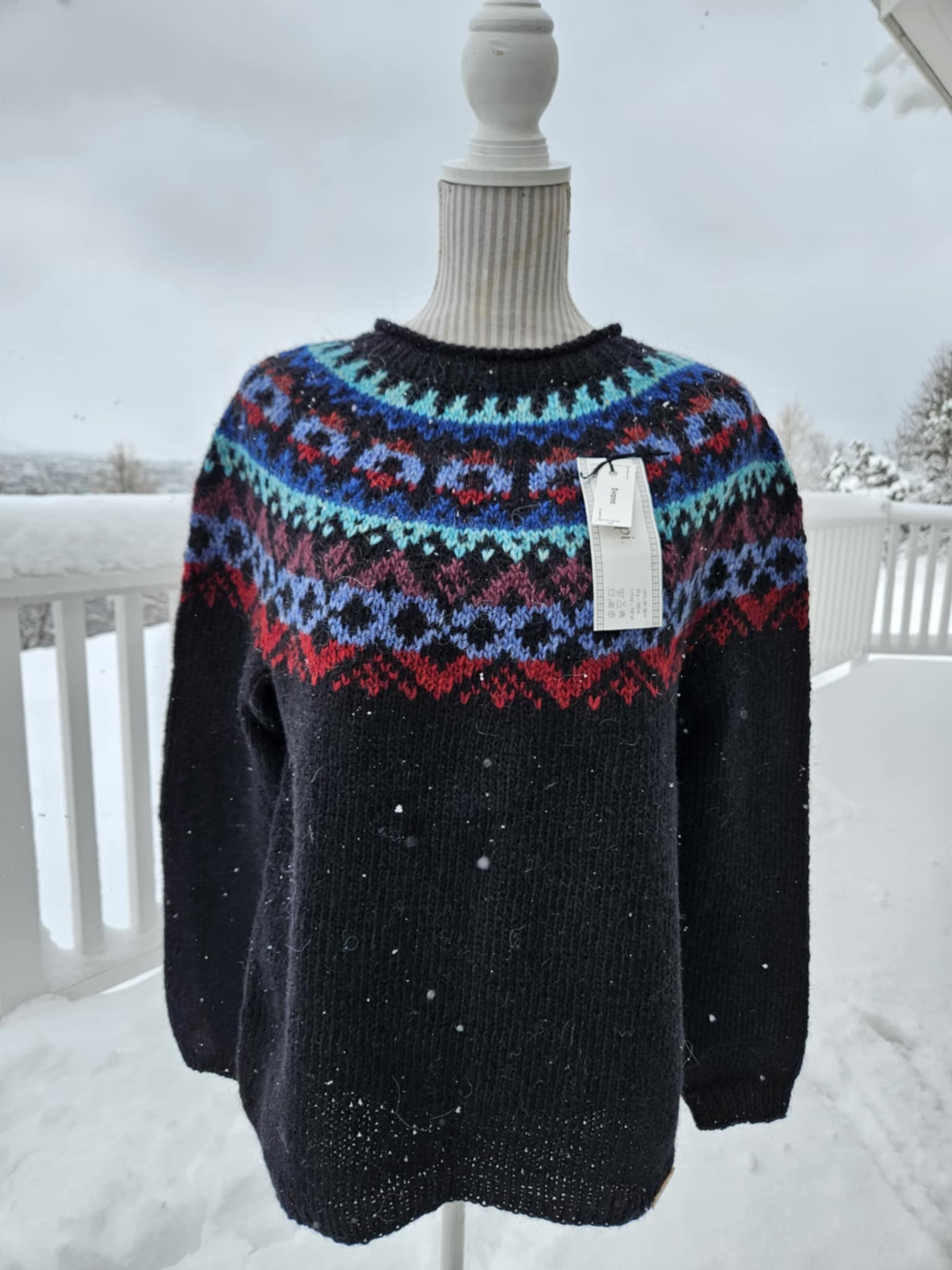 A black Icelandic wool sweater with a colorful geometric pattern on the yoke, displayed on a mannequin against a snowy backdrop.