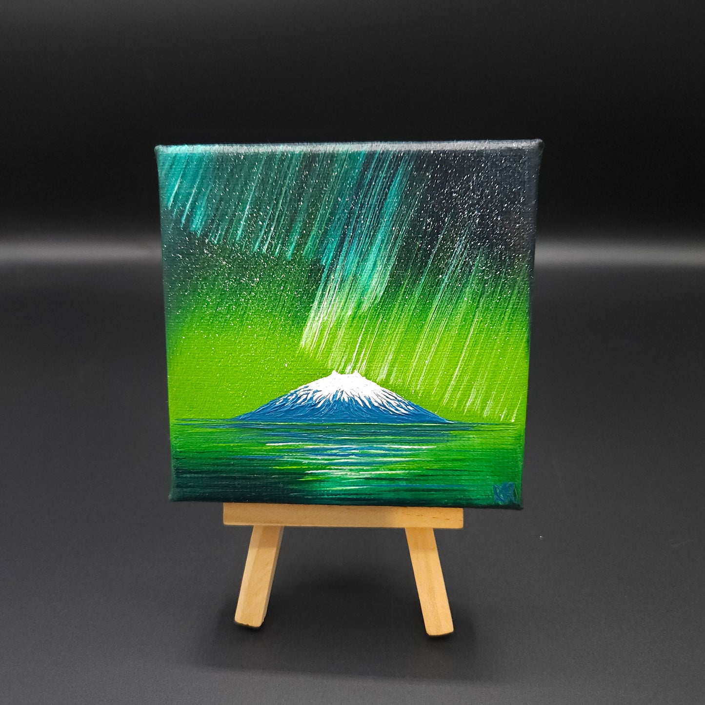 Northern lights landscape painting featuring blue and green northern lights over a snow-capped mountain reflected in water.