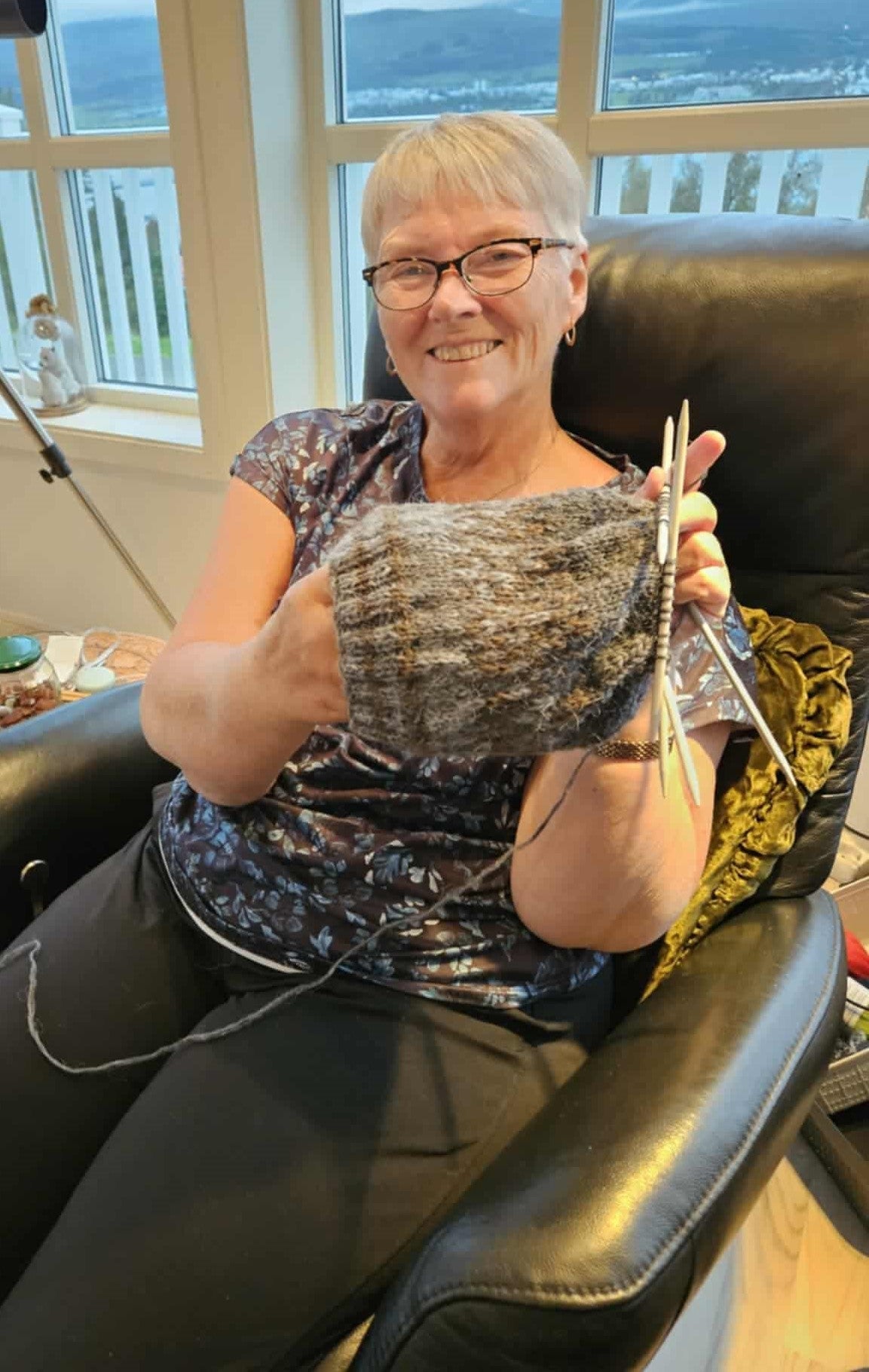A woman, smiling and holding up her knitwork. Sitting in a chair.