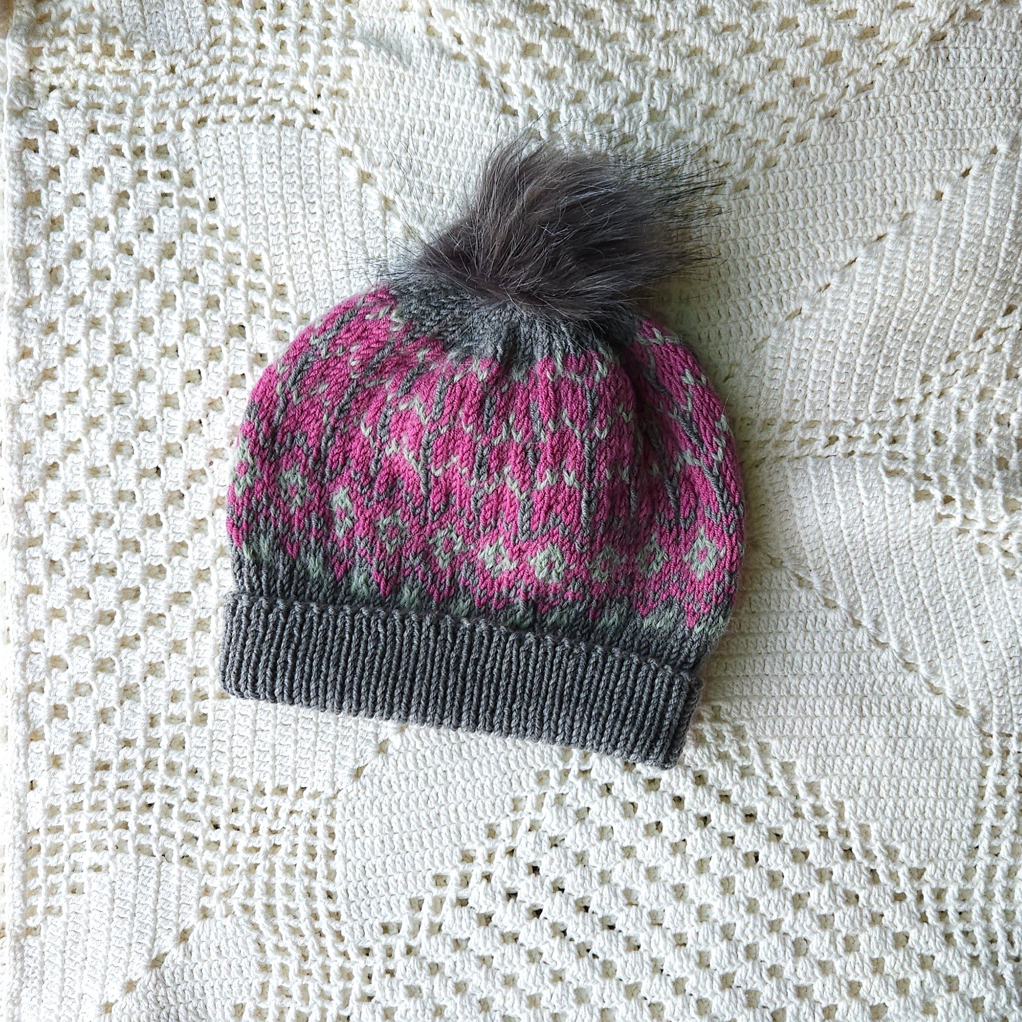 Knitted hat with a pink pattern and gray pom-pom on a textured white background
