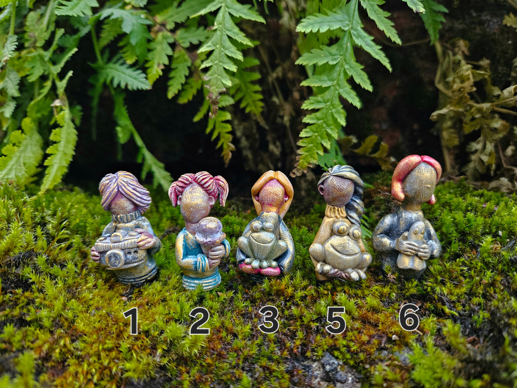 Set of six small figurines with different hairstyles on a mossy surface with ferns in the background.