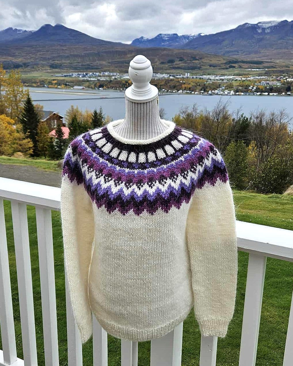 Knitted sweater with a colorful pattern on a mannequin against a scenic backdrop of mountains and a lake.