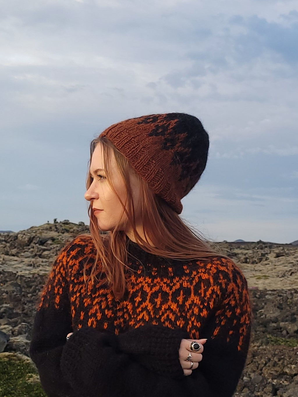 Woman wearing a patterned sweater and knit hat standing in a rocky landscape with a cloudy sky.