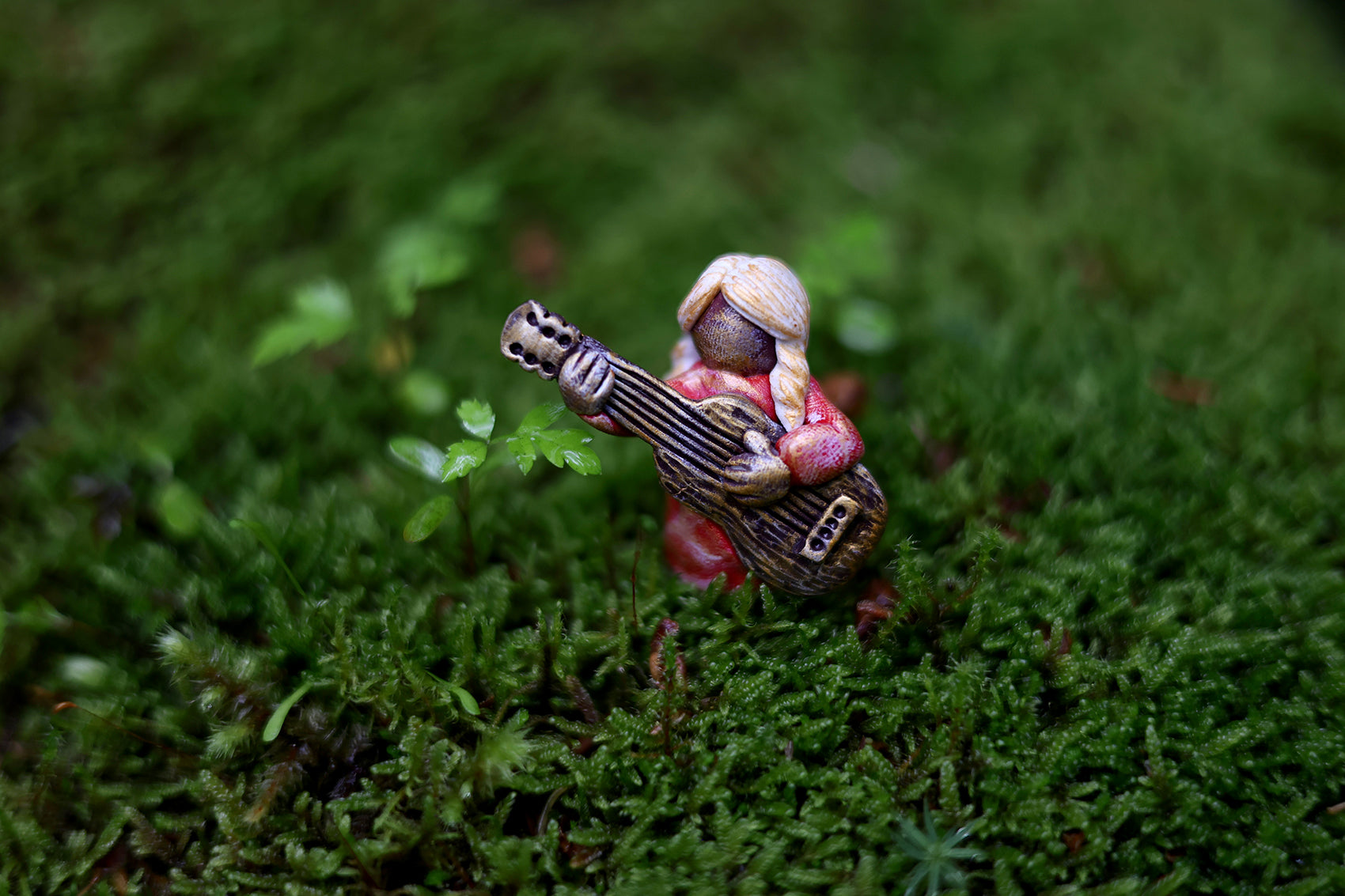 A miniature polymer clay figure depicting a blond person playing a guitar, surrounded by green moss.