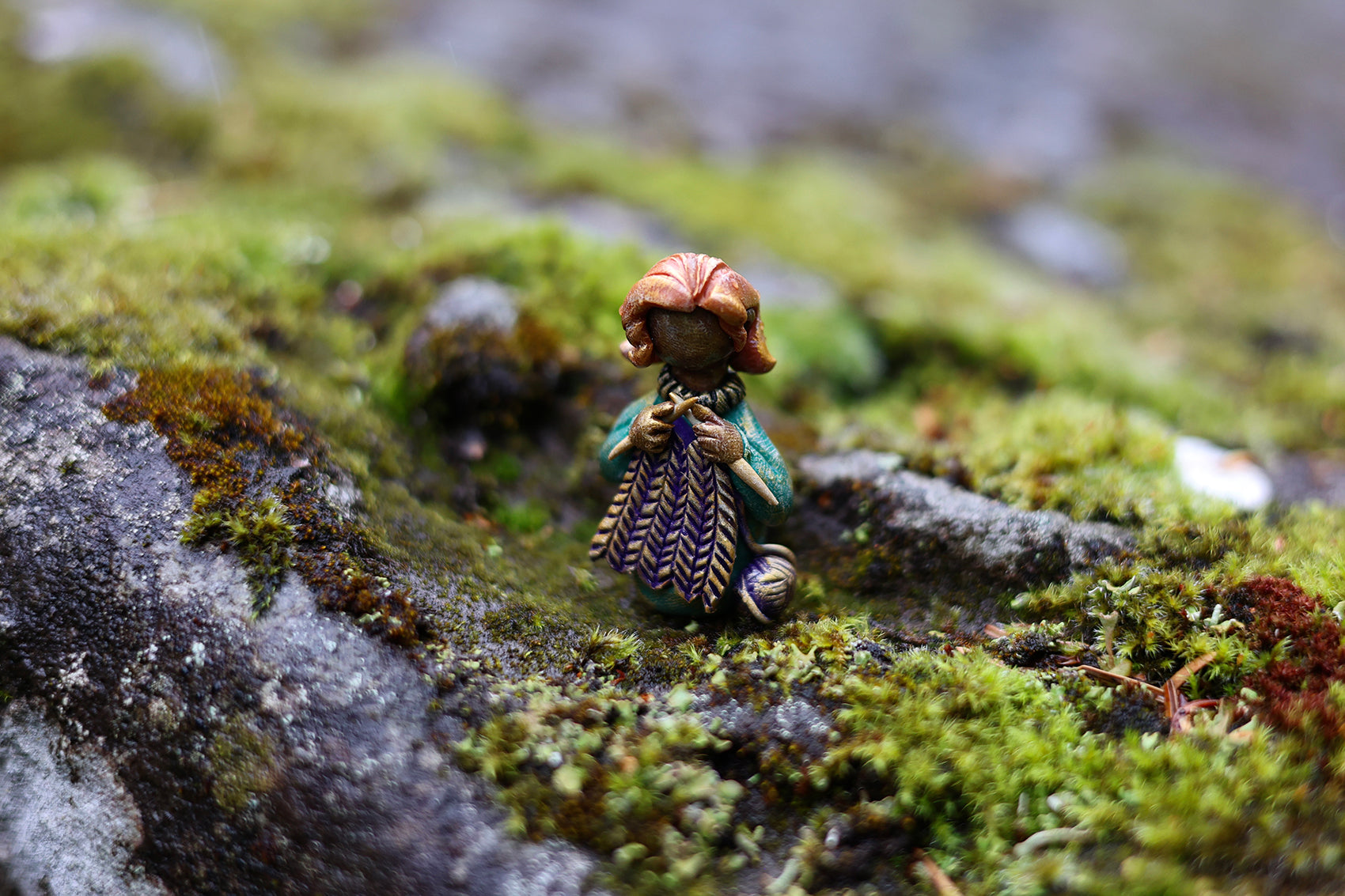 A miniature polymer clay figure depicting a homemaker, placed on a mossy rock surface.