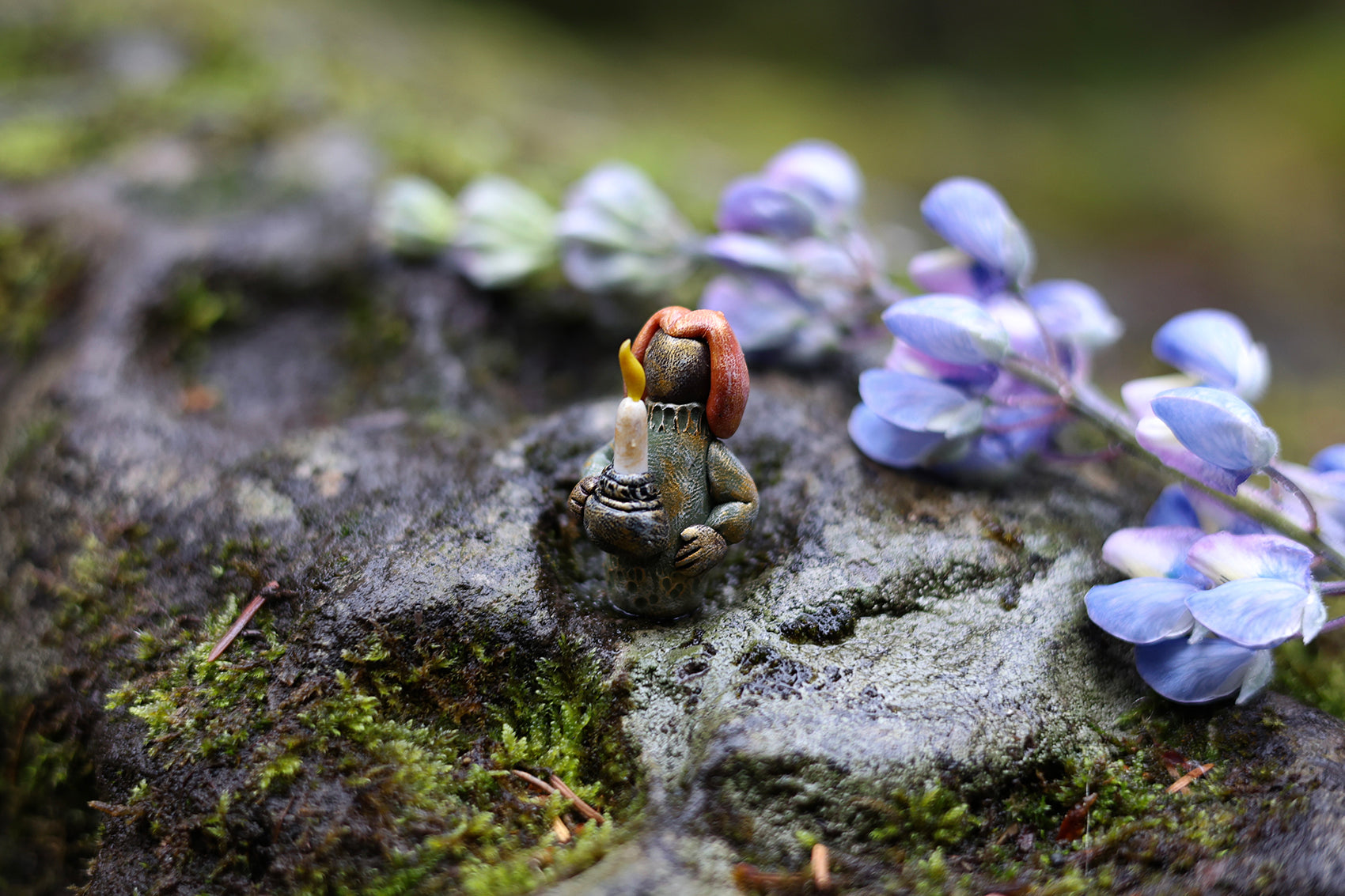 Miniature polymer clay figure depicting a scene with a hidden person holding a candle, surrounded by purple flowers and green moss on a rocky surface.