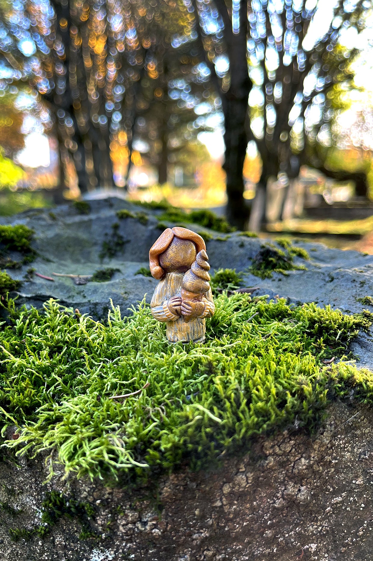 A miniature polymer clay figure depicting a hidden person sitting on a mossy rock, surrounded by trees in the background.