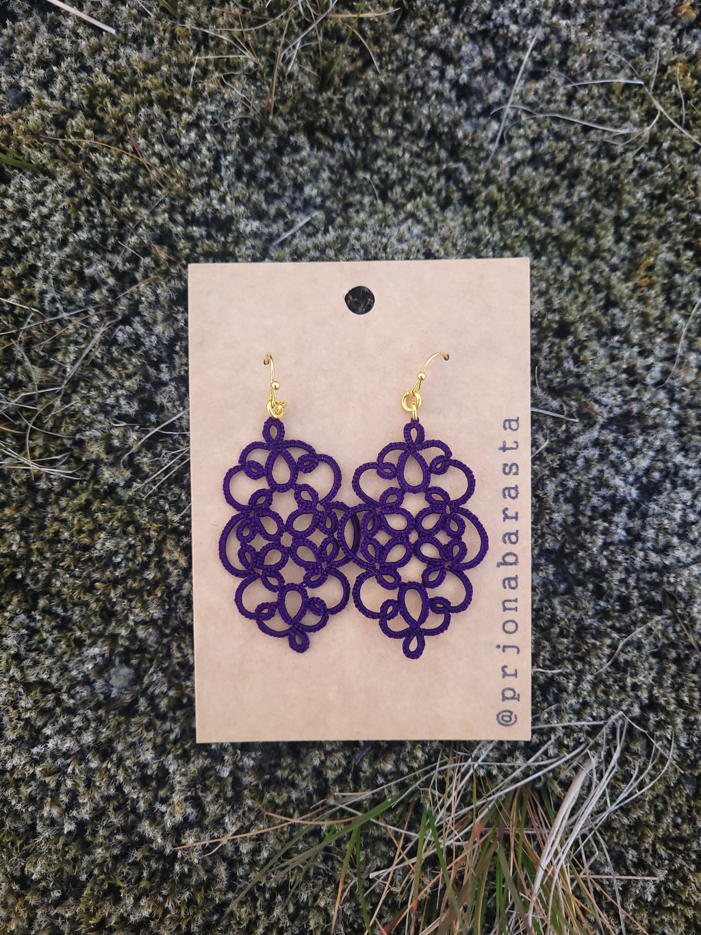 Purple lace earrings on a beige card with 'prijonabarasta' branding, placed on a textured green surface.