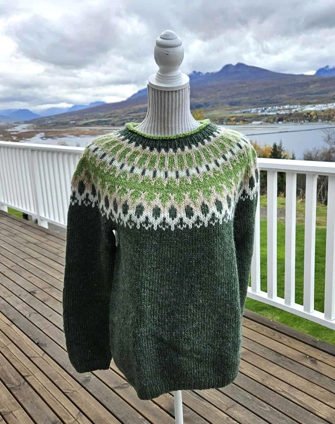 Green knitted sweater with a patterned yoke on a mannequin against a scenic background.