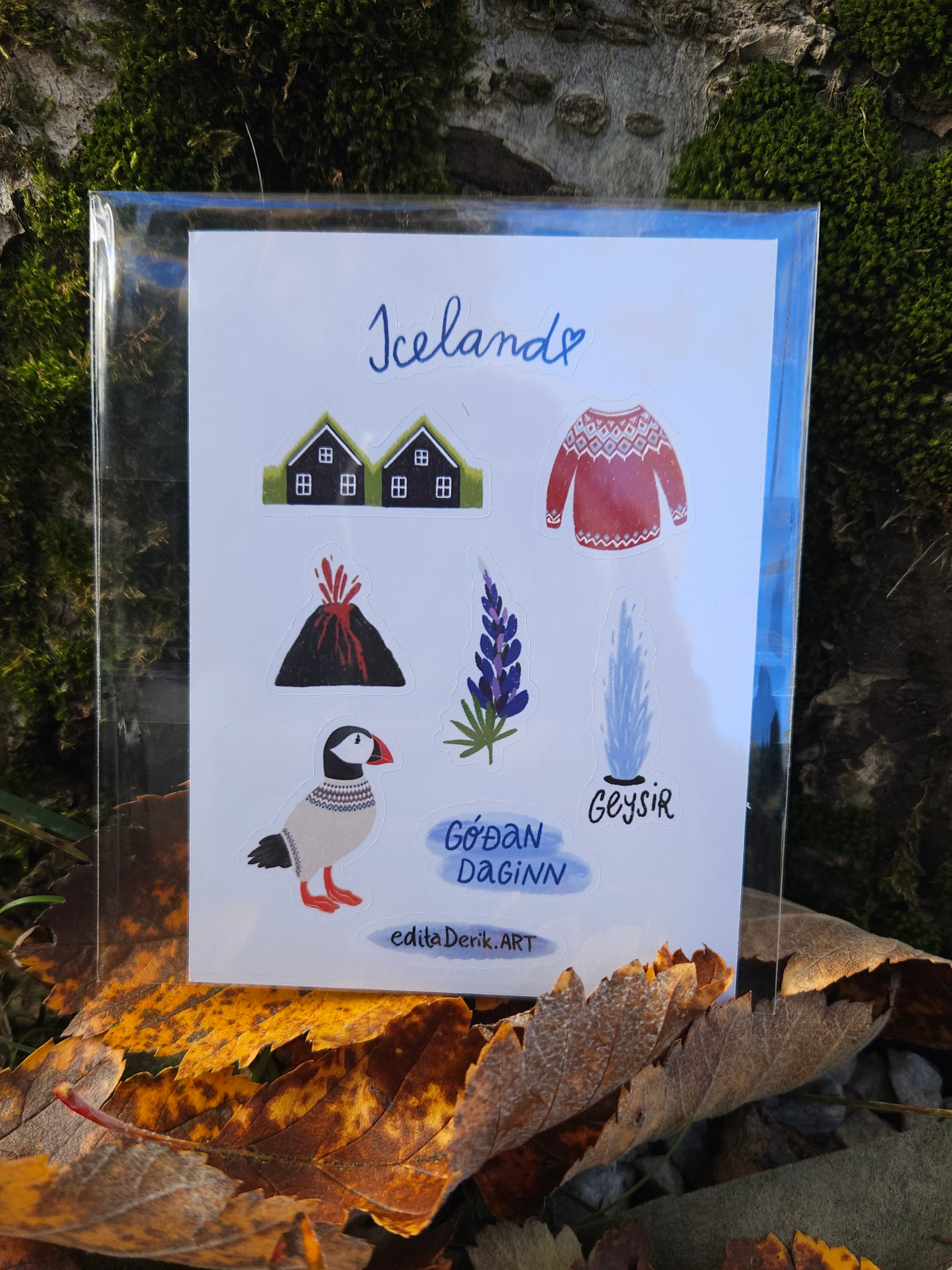 Card with illustrations of Icelandic elements like houses, a sweater, mountains, a puffin, and a geyser, placed outdoors.