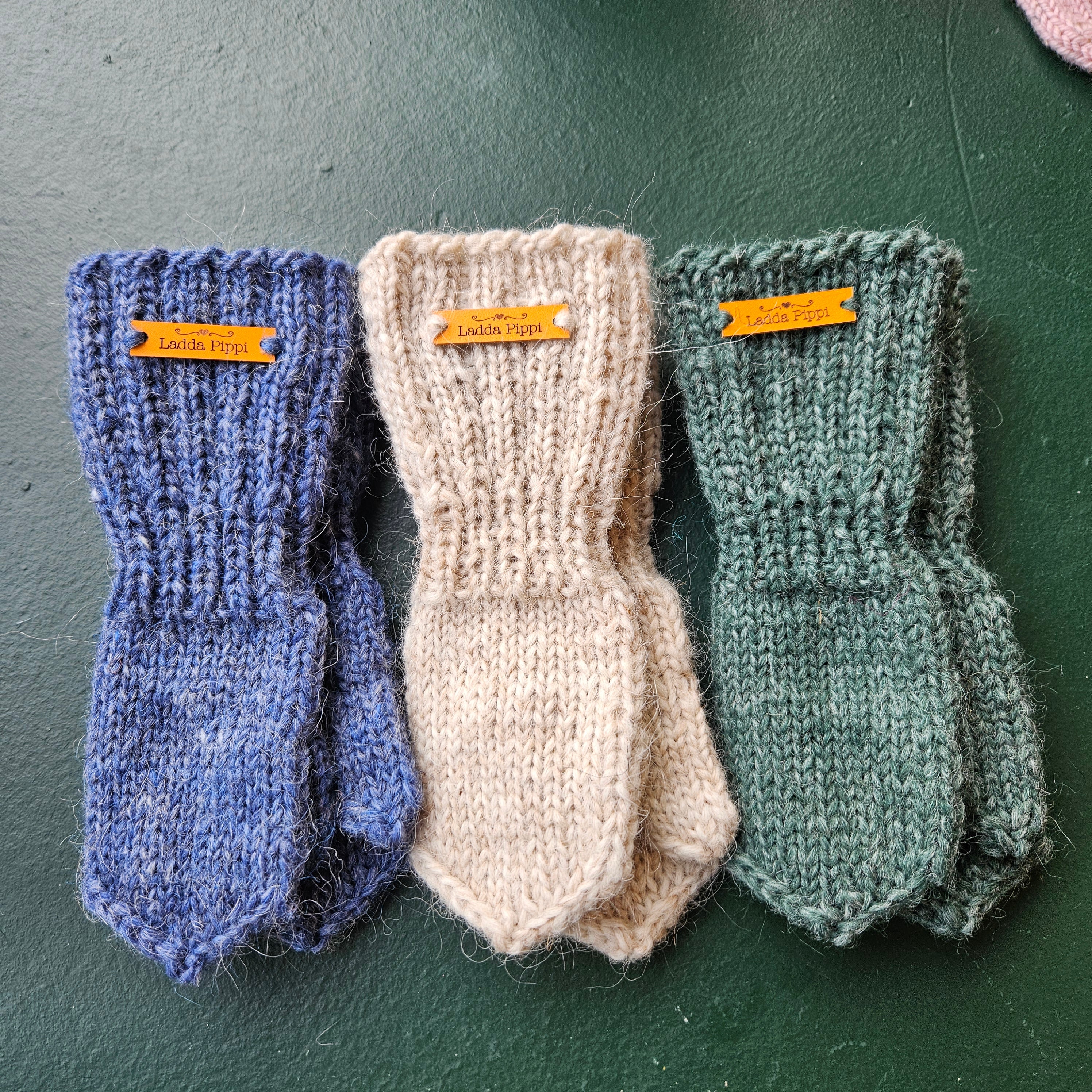 Three pairs of knitted mittens in green, beige, and blue on a dark surface.