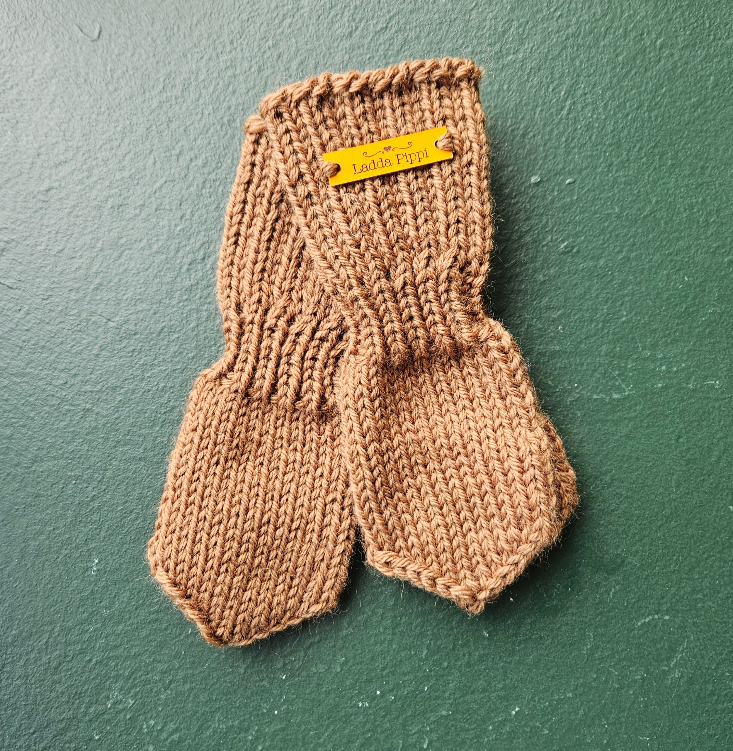 Pair of brown knitted baby mittens with a yellow label on a green background