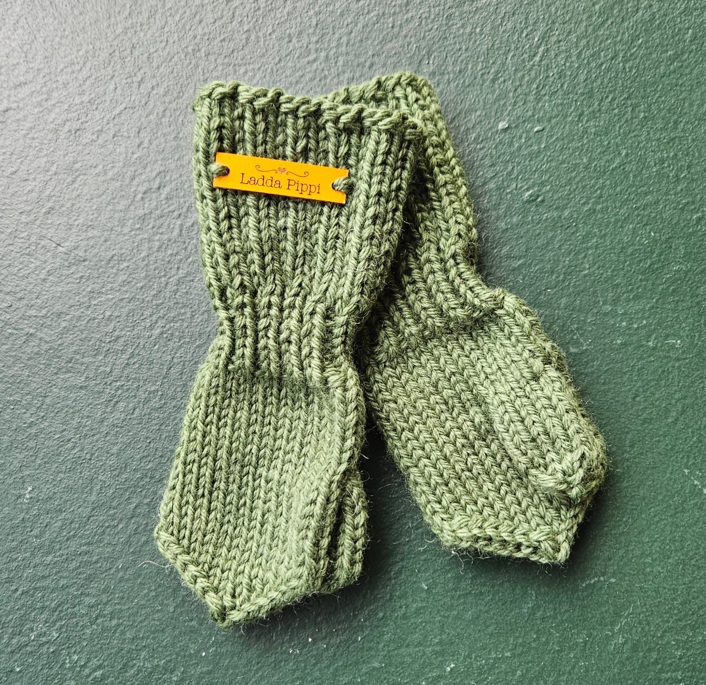 Pair of green knitted baby mittens with a brand tag on a green background