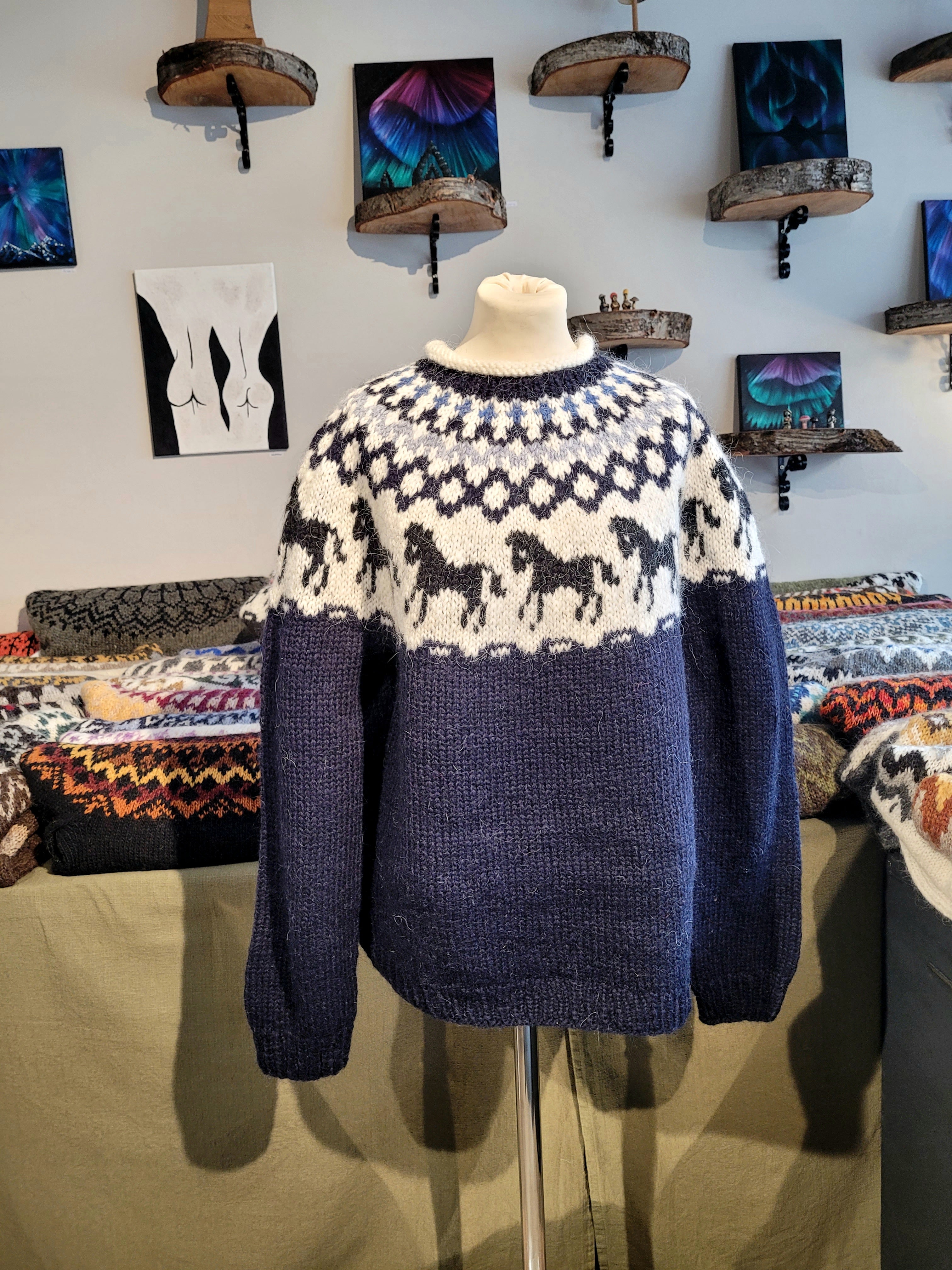 A mannequin in a blue hand-knit Icelandic wool sweater featuring a traditional Nordic pattern in white and light blue colors with black horses in a store setting.