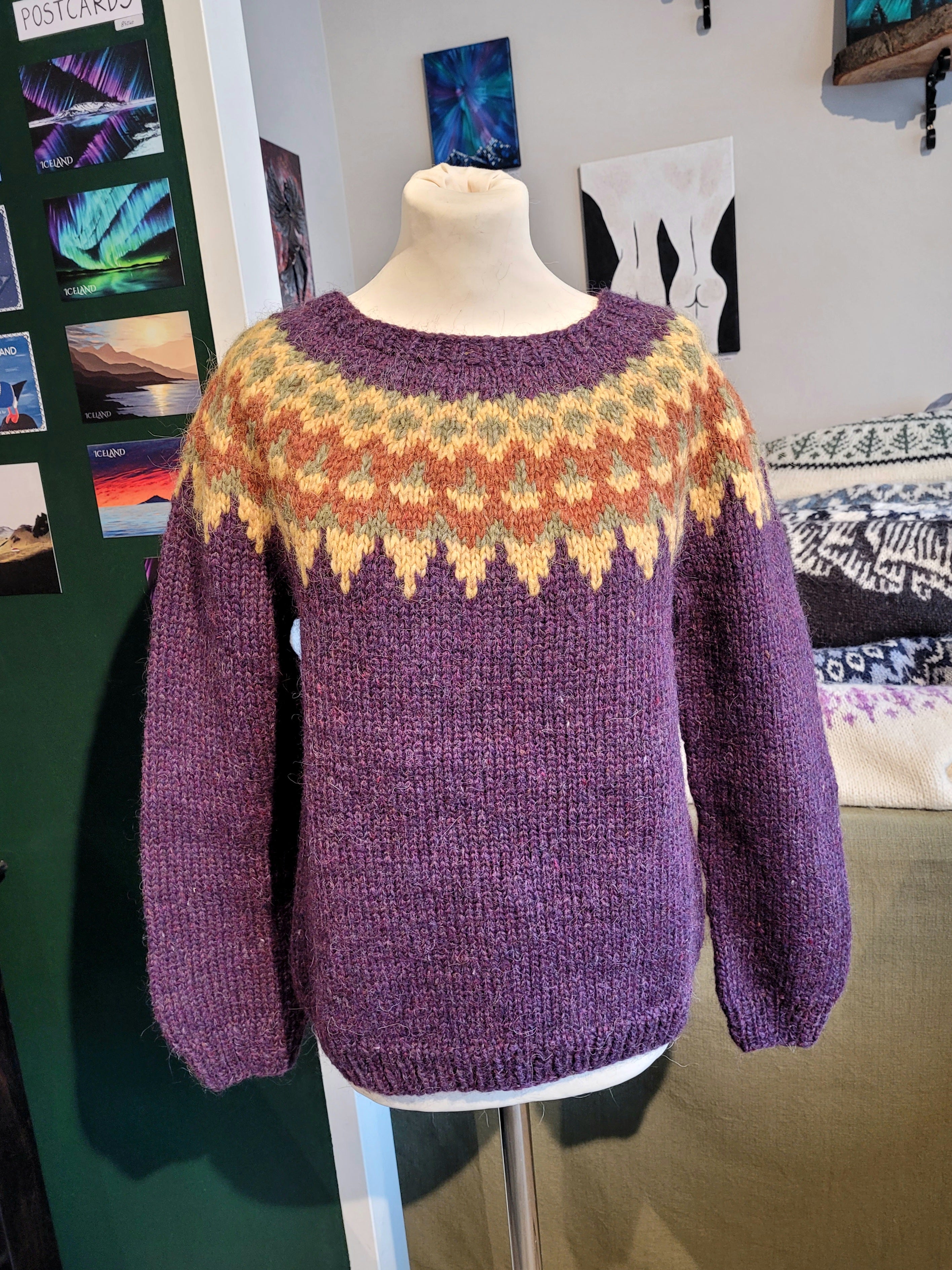 A mannequin in a purple hand-knit Icelandic wool sweater featuring a traditional Nordic pattern in brown and yellow colors in a store setting.