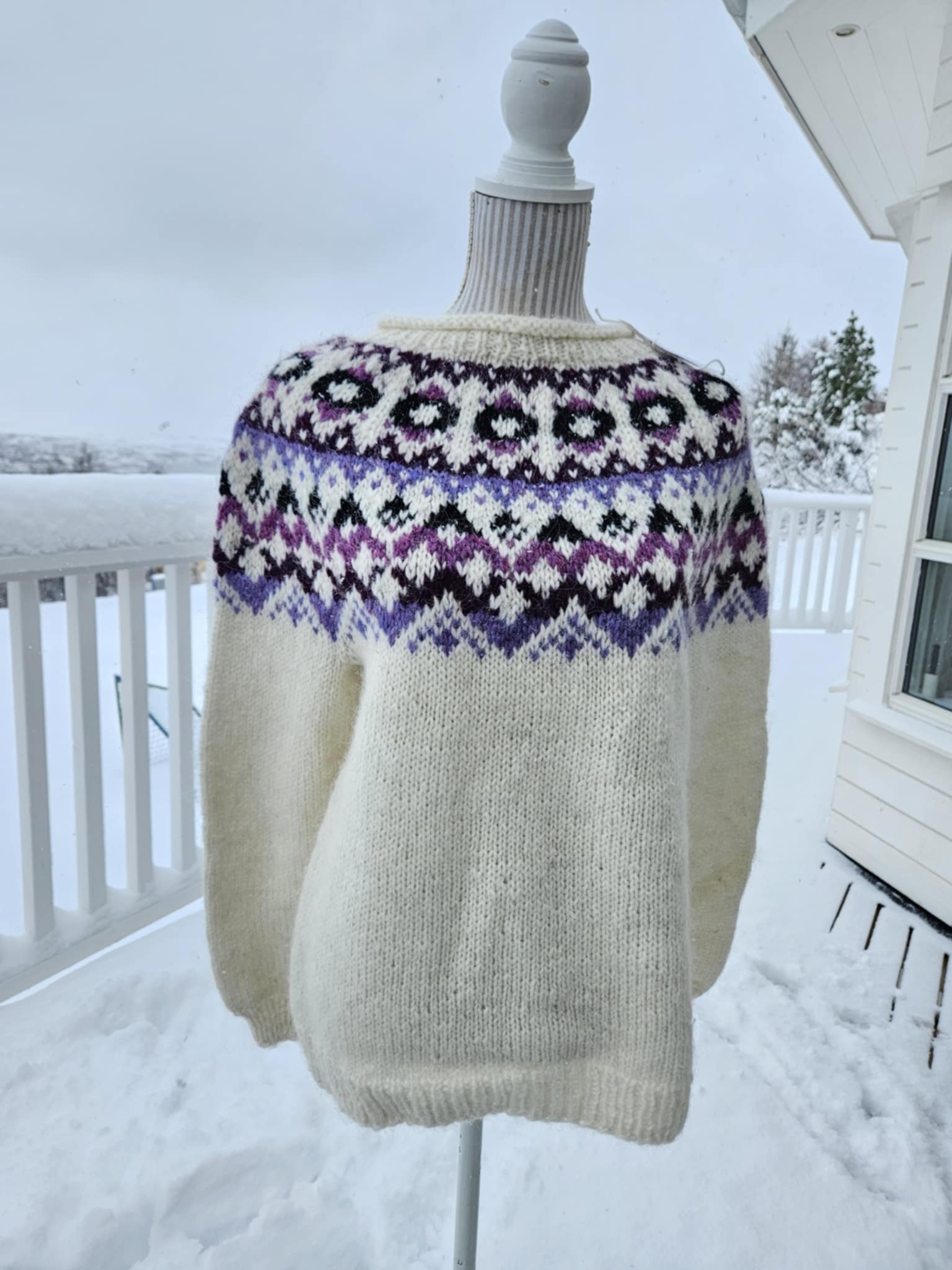 A hand knit Icelandic wool sweater with a traditional Nordic pattern in purple and black colors on a mannequin outdoors in a snowy setting.