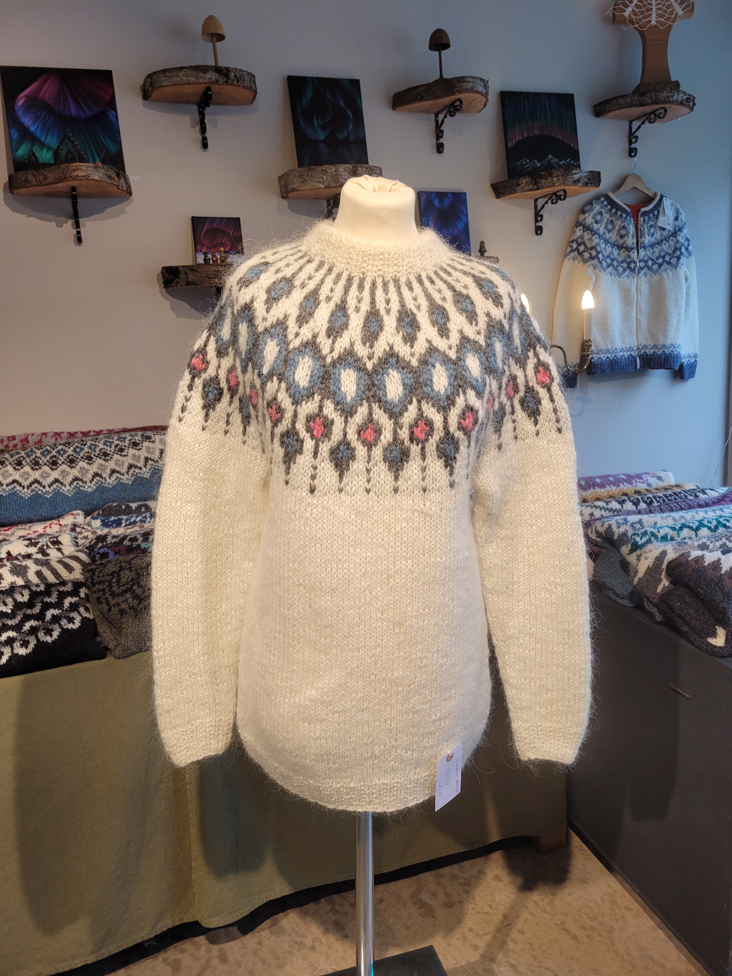 A mannequin in a white hand-knit Icelandic wool sweater featuring a traditional Nordic pattern in pink and blue colors in a store setting.