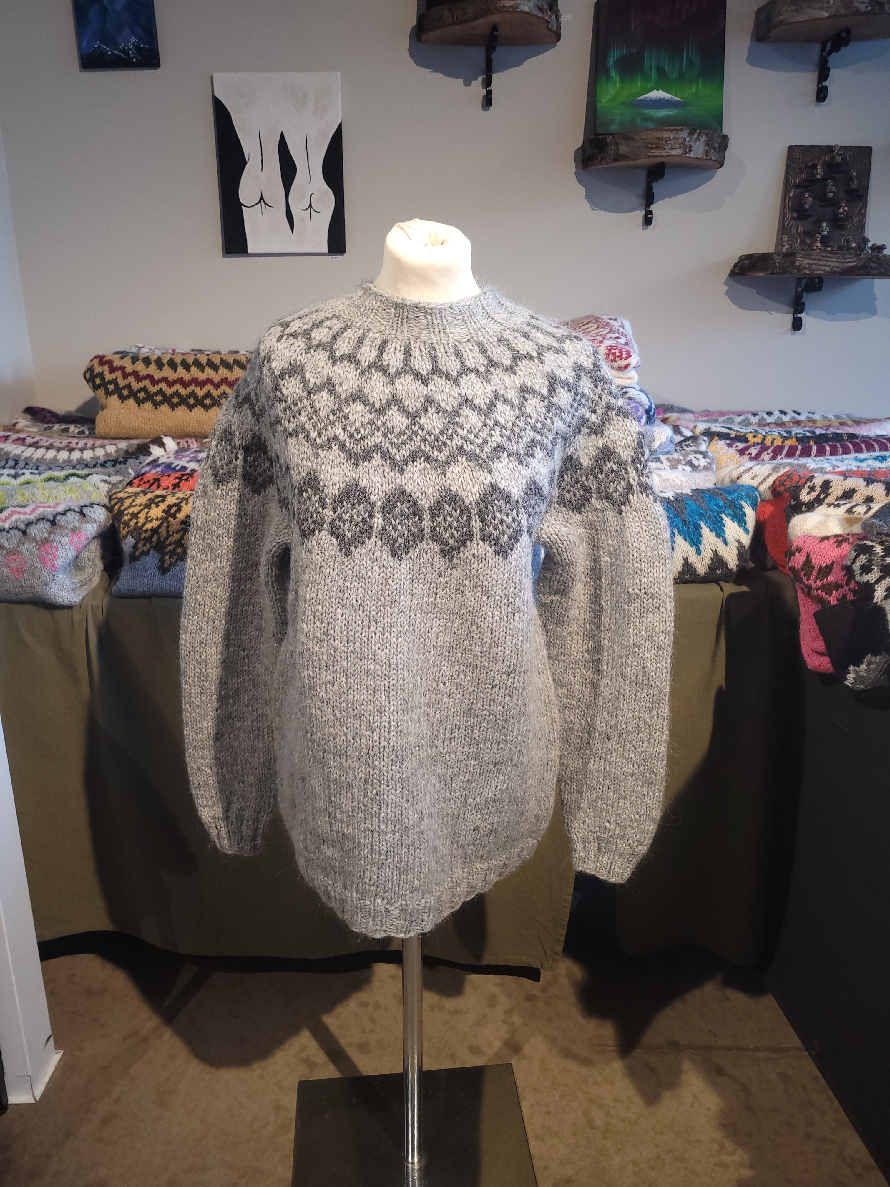 A mannequin in a light gray hand-knit Icelandic wool sweater featuring a traditional Nordic pattern in dark gray in a store setting.