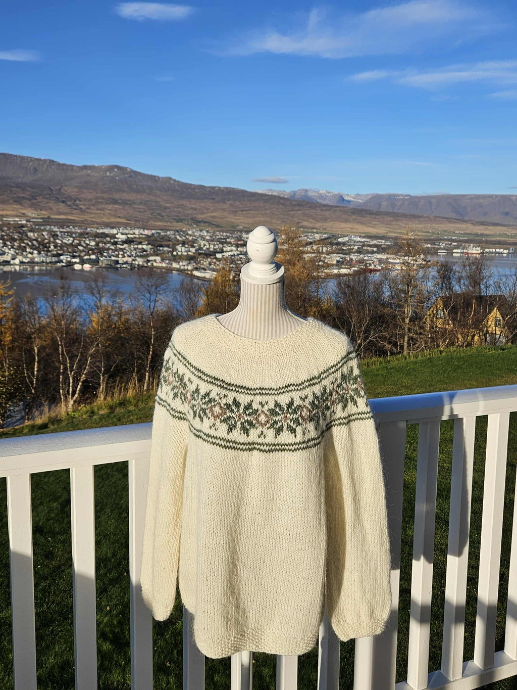 White-colored sweater with green patterns on a mannequin against a scenic backdrop of mountains and water.