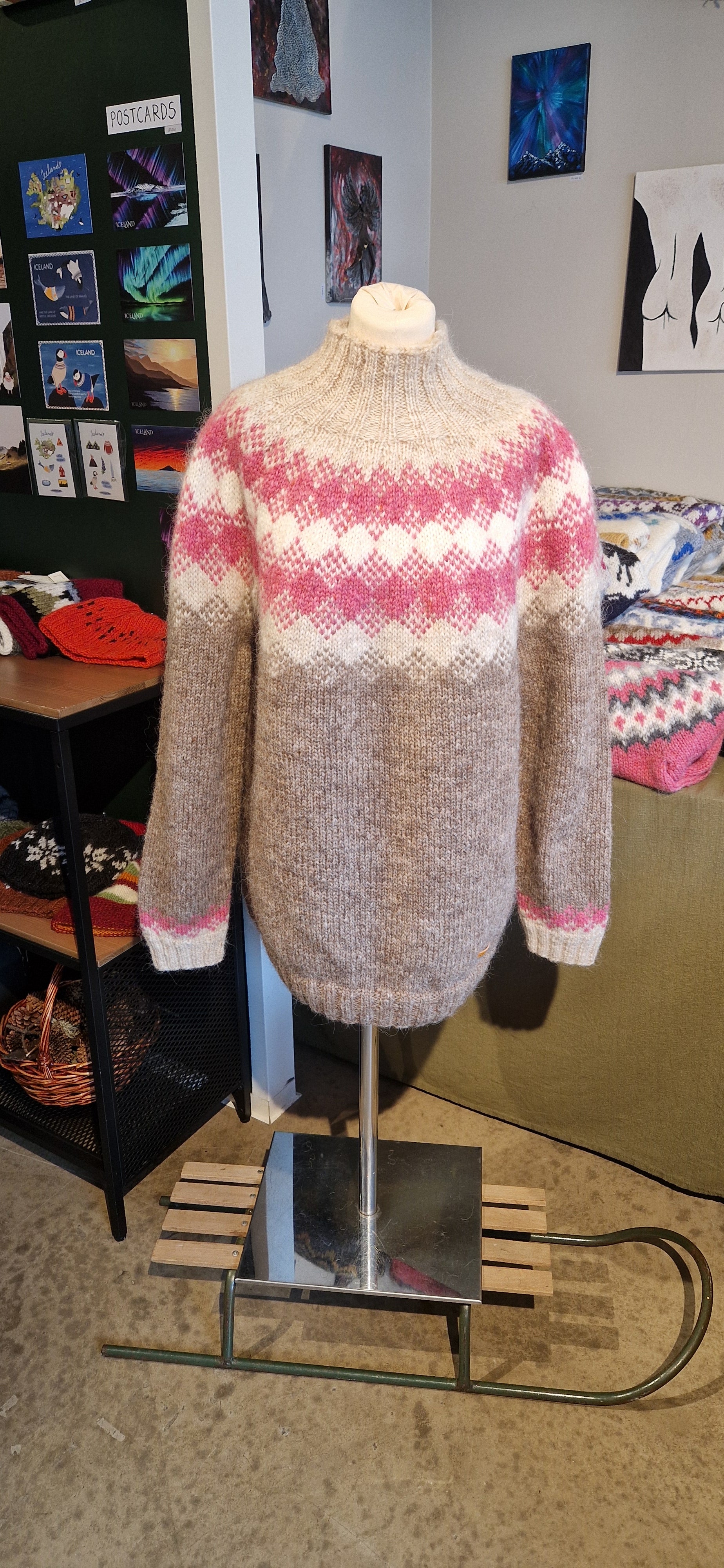 A mannequin in a beige hand-knit Icelandic wool sweater featuring a traditional Nordic pattern in pink and white in a store setting.