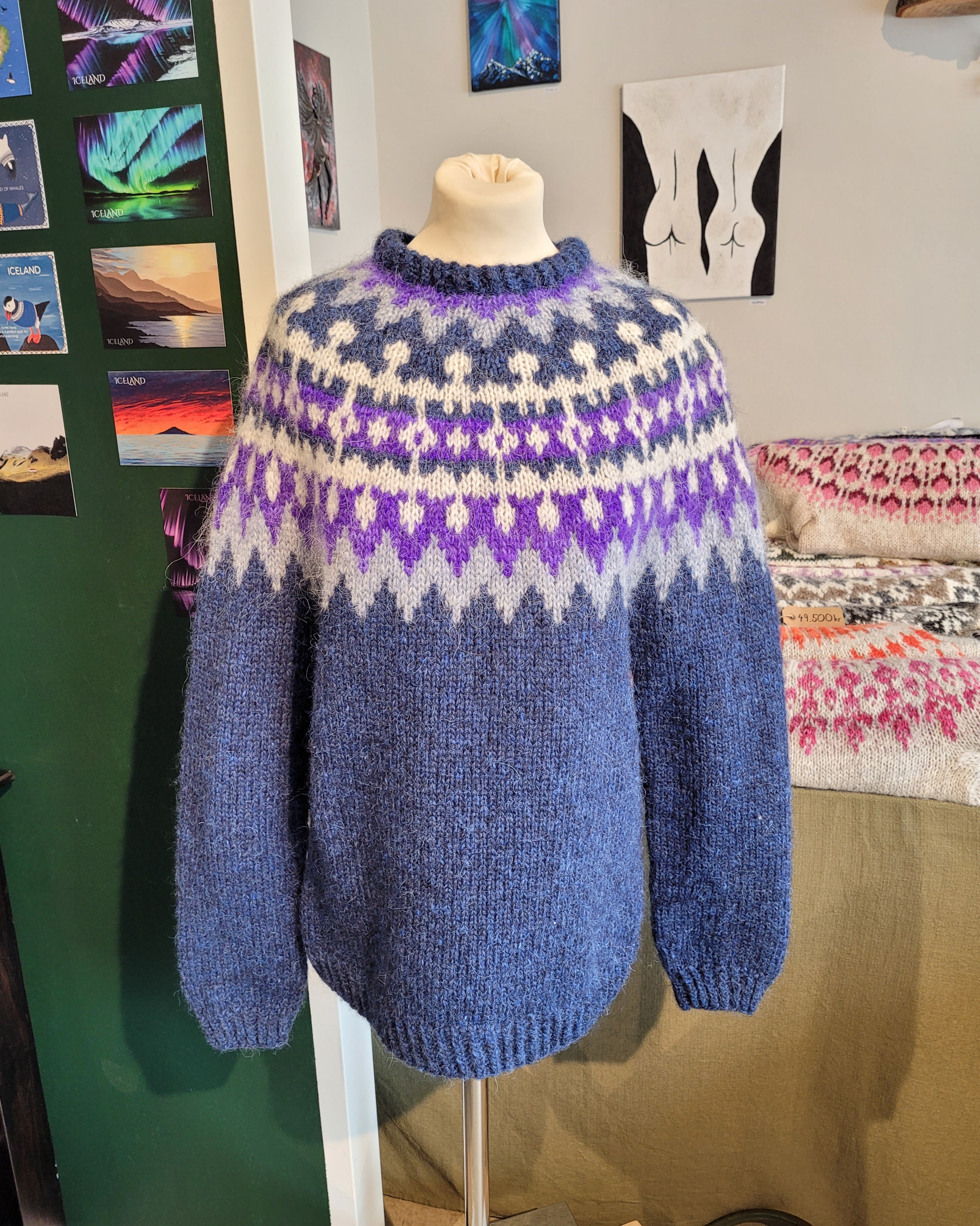 A mannequin in a blue hand-knit Icelandic wool sweater featuring a traditional Nordic pattern in white and purple in a store setting.