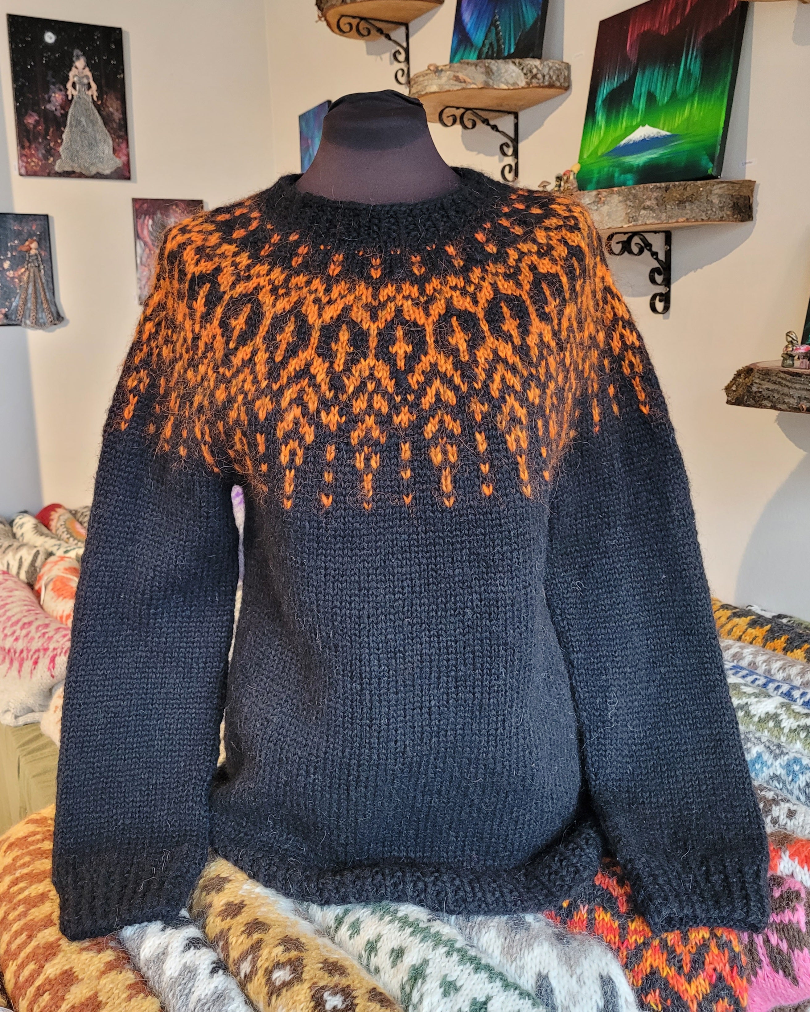 A mannequin in a black hand-knit Icelandic wool sweater featuring a traditional Nordic pattern in orange in a store setting.
