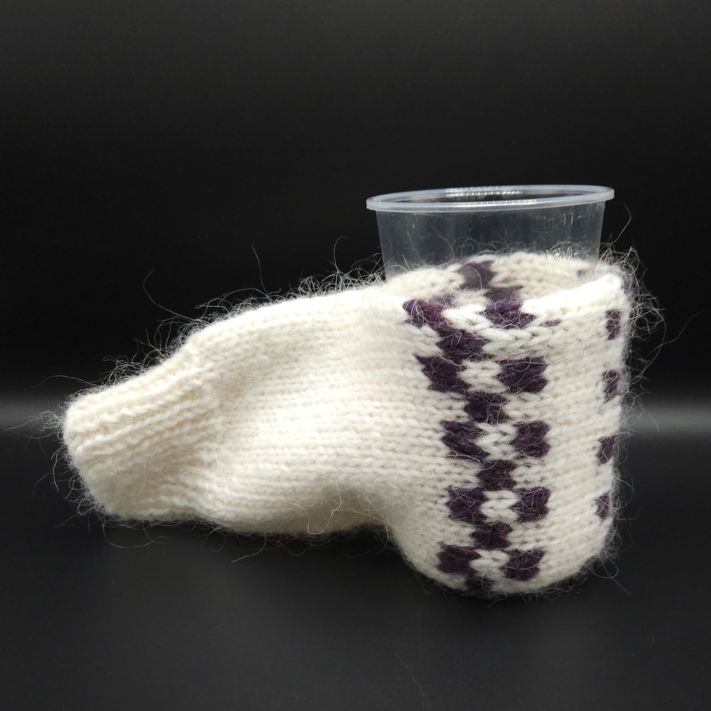 Knitted white and purple mitten holding a clear plastic cup on a black background