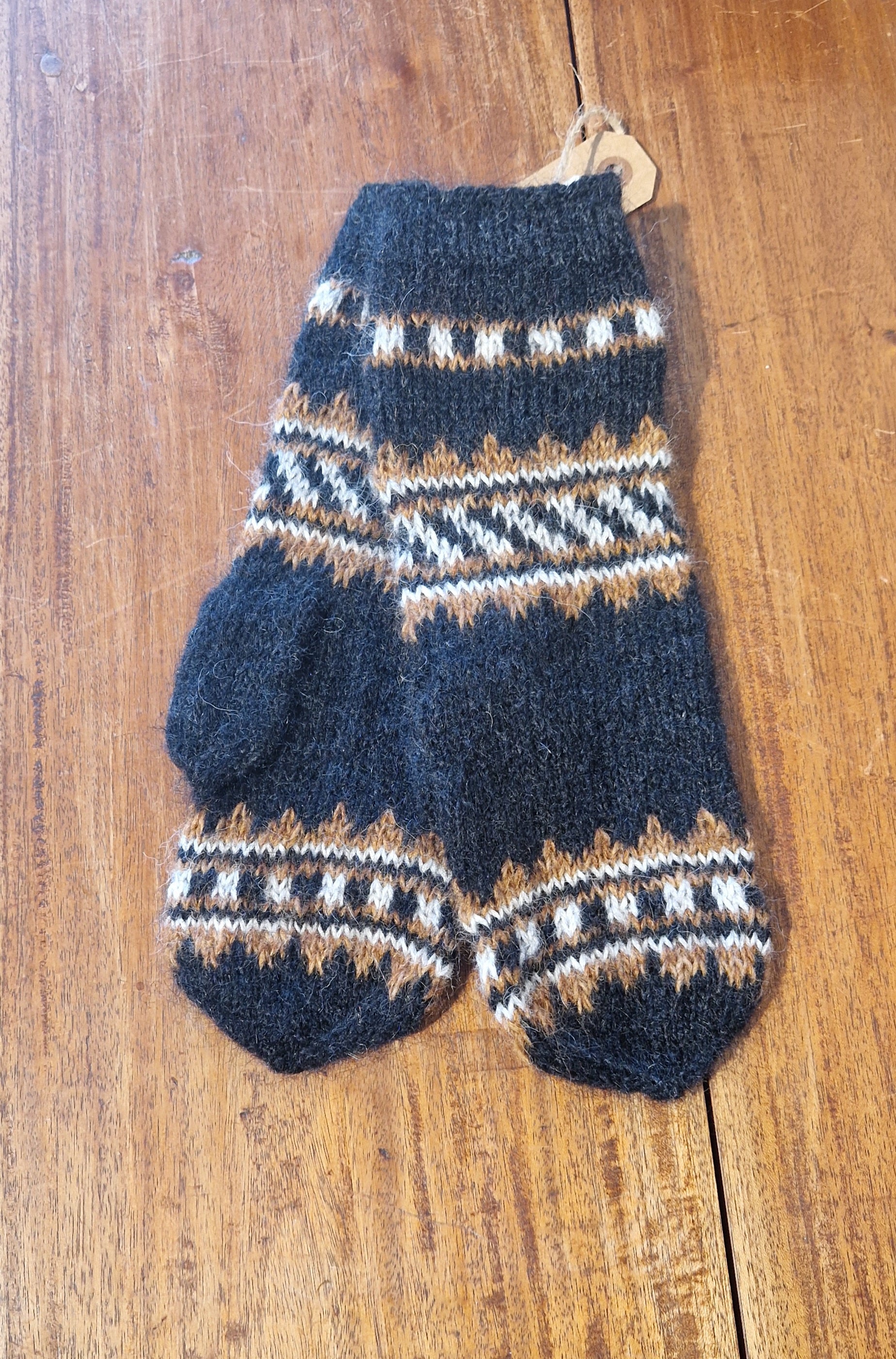 A pair of black knitted wool mittens with a geometric pattern in beige and white colors, displayed on a wooden surface.