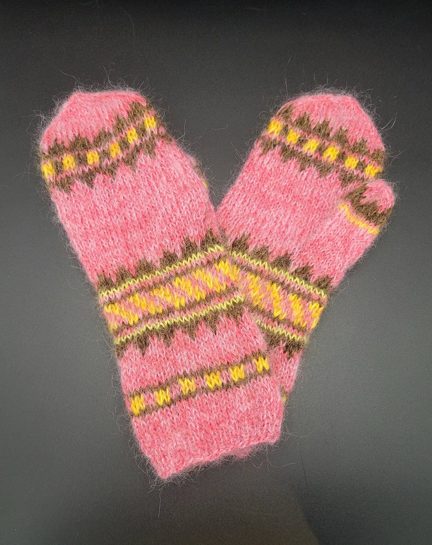 Pair of pink knitted mittens with yellow and brown patterns on a dark background