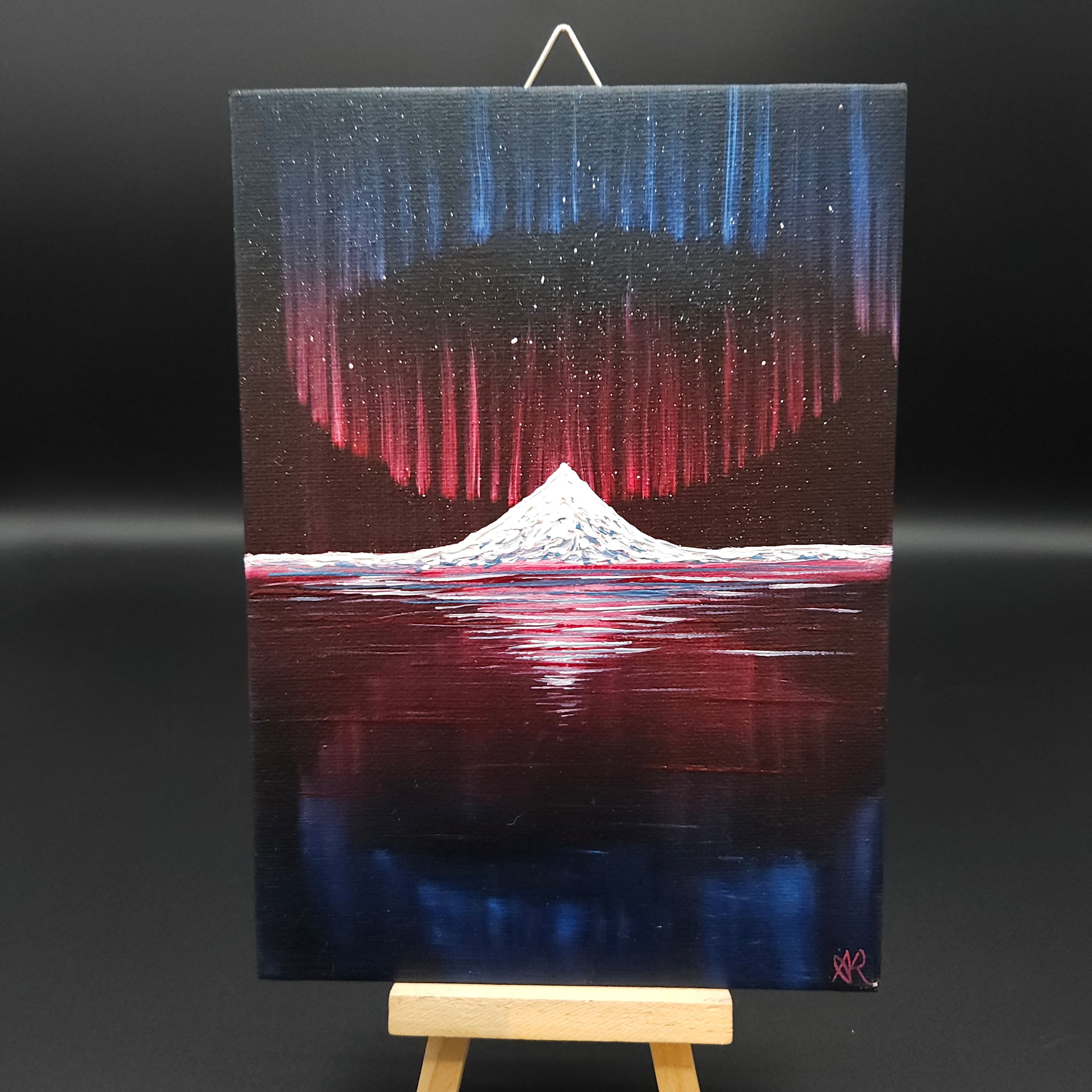 Northern lights landscape painting featuring blue and red northern lights over a snow-capped mountain reflected in water.