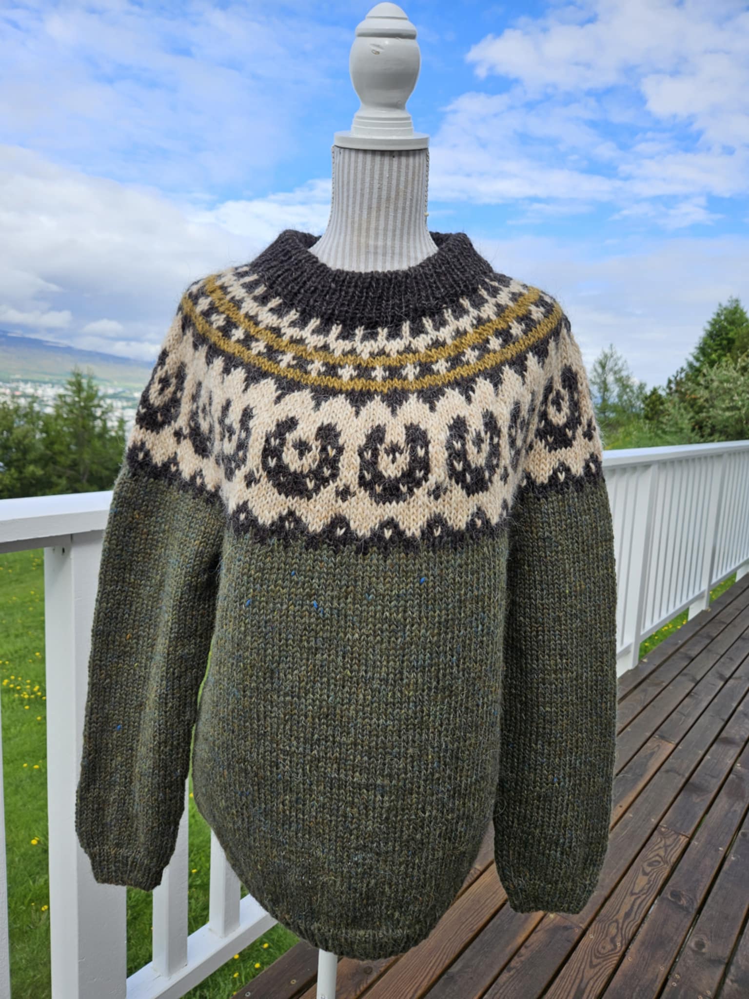 Green sweater with patterned neckline on a mannequin against a scenic outdoor background