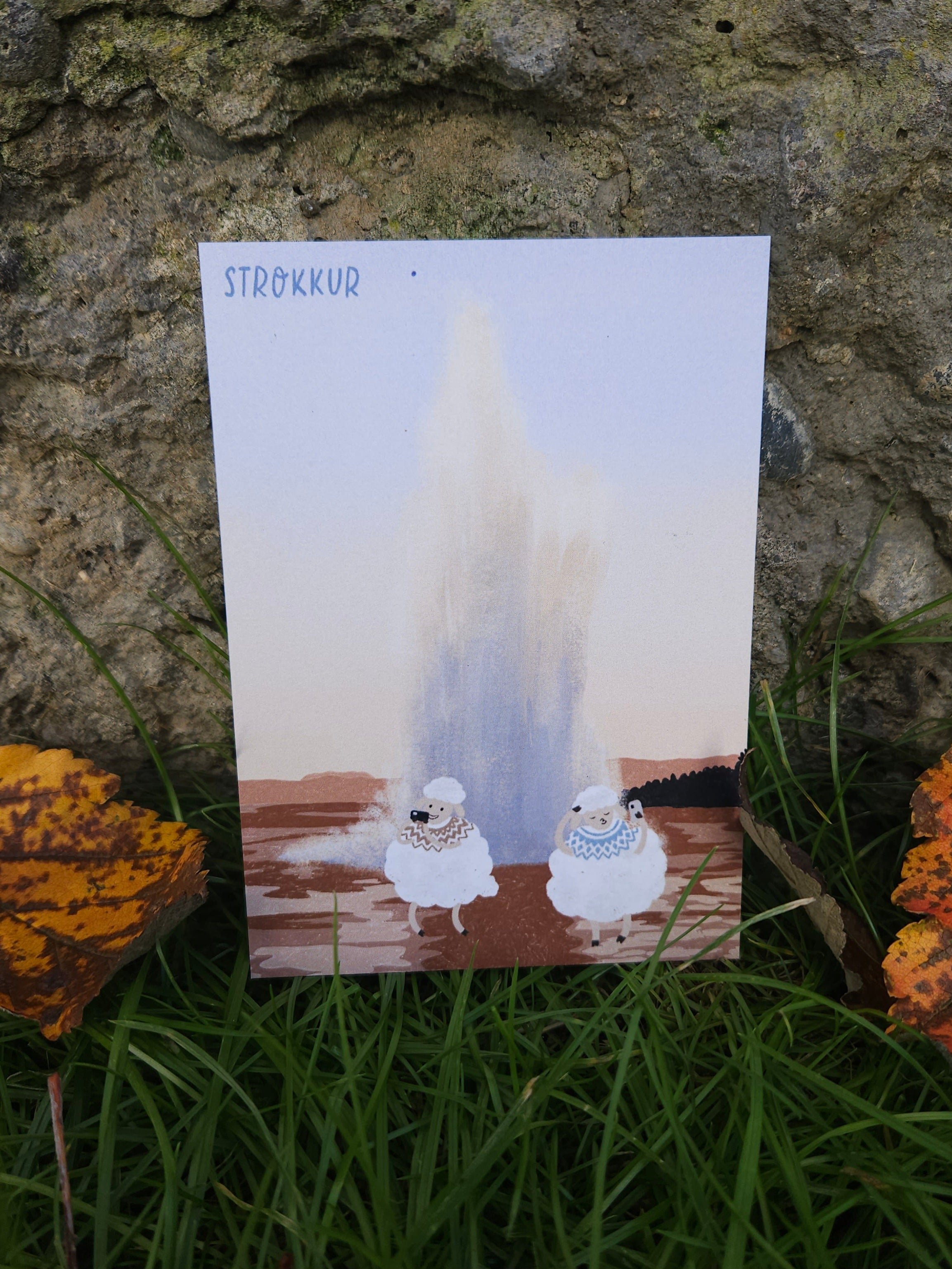 Postcard with two cartoon sheep in front of a geysir on a stone surface with grass and leaves.