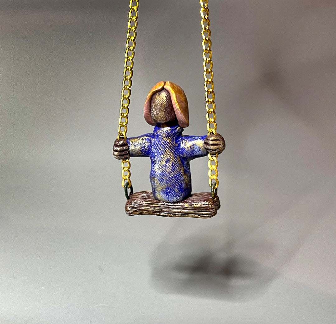 Small figurine of a person on a swing with a gold chain against a gray background