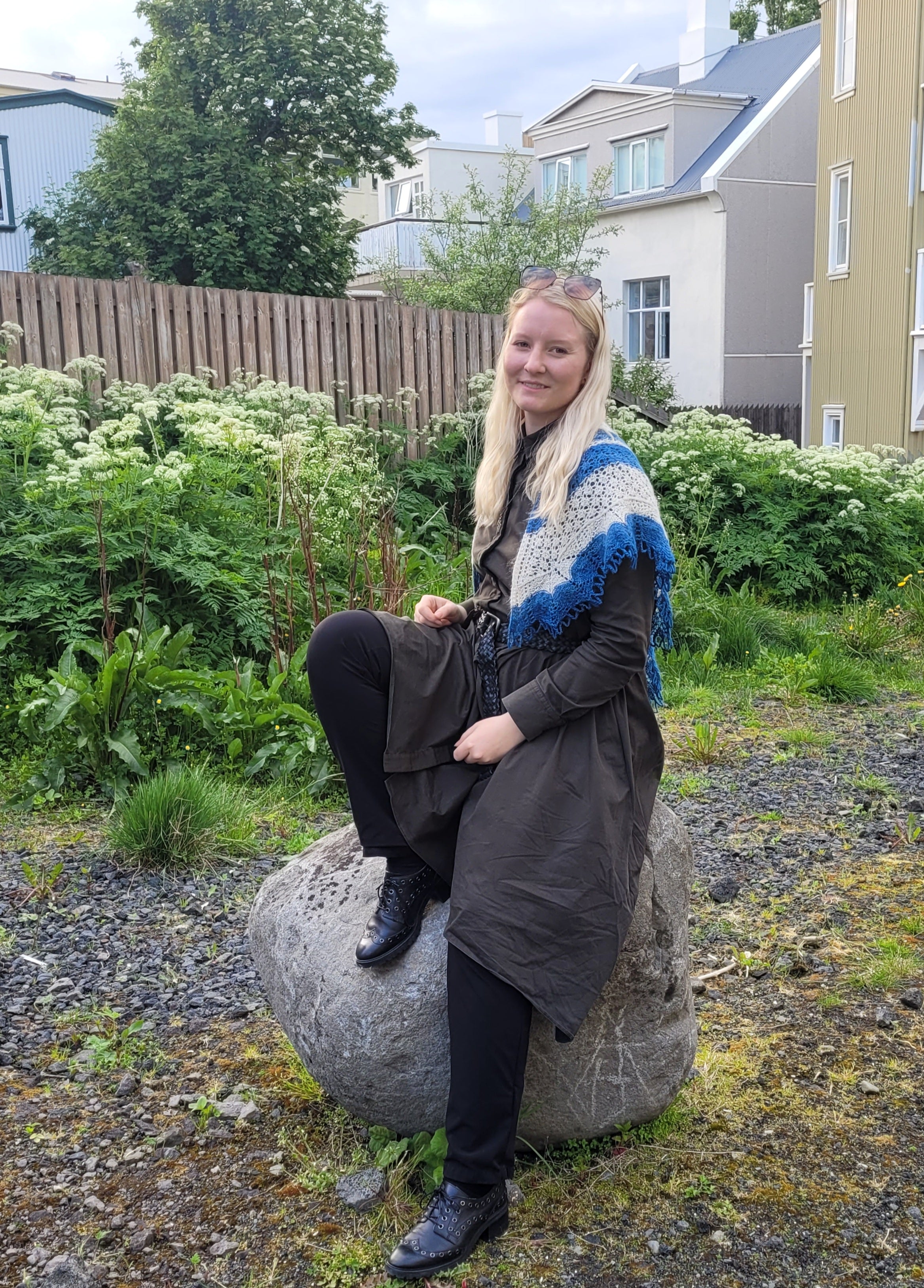 Woman sitting on a rock in a garden with houses in the background with a white and blue shawl on her shoulders.
