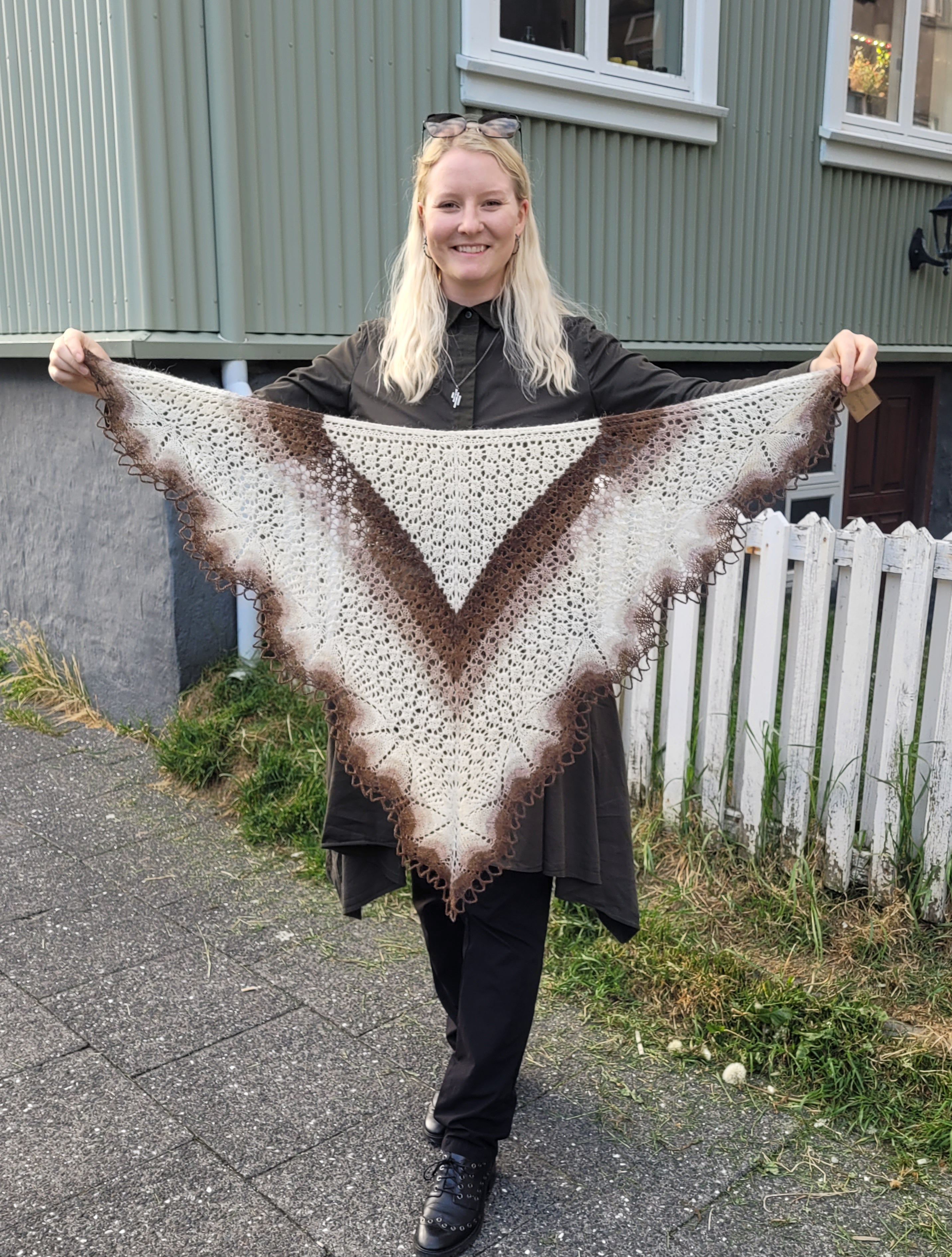 Person holding a large, triangular, brown and white knitted shawl outdoors.