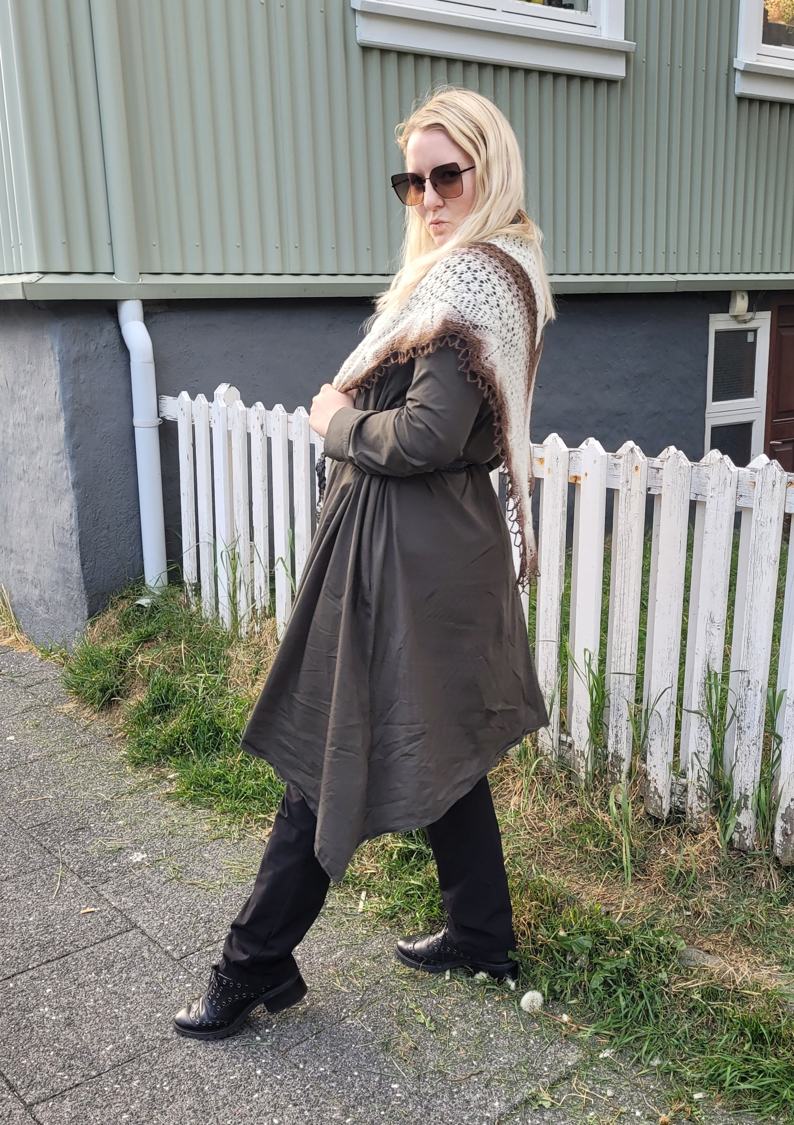 Woman wearing a long coat and sunglasses standing on a sidewalk with a building and white picket fence in the background with a brown and white knitted shawl on her shoulders.