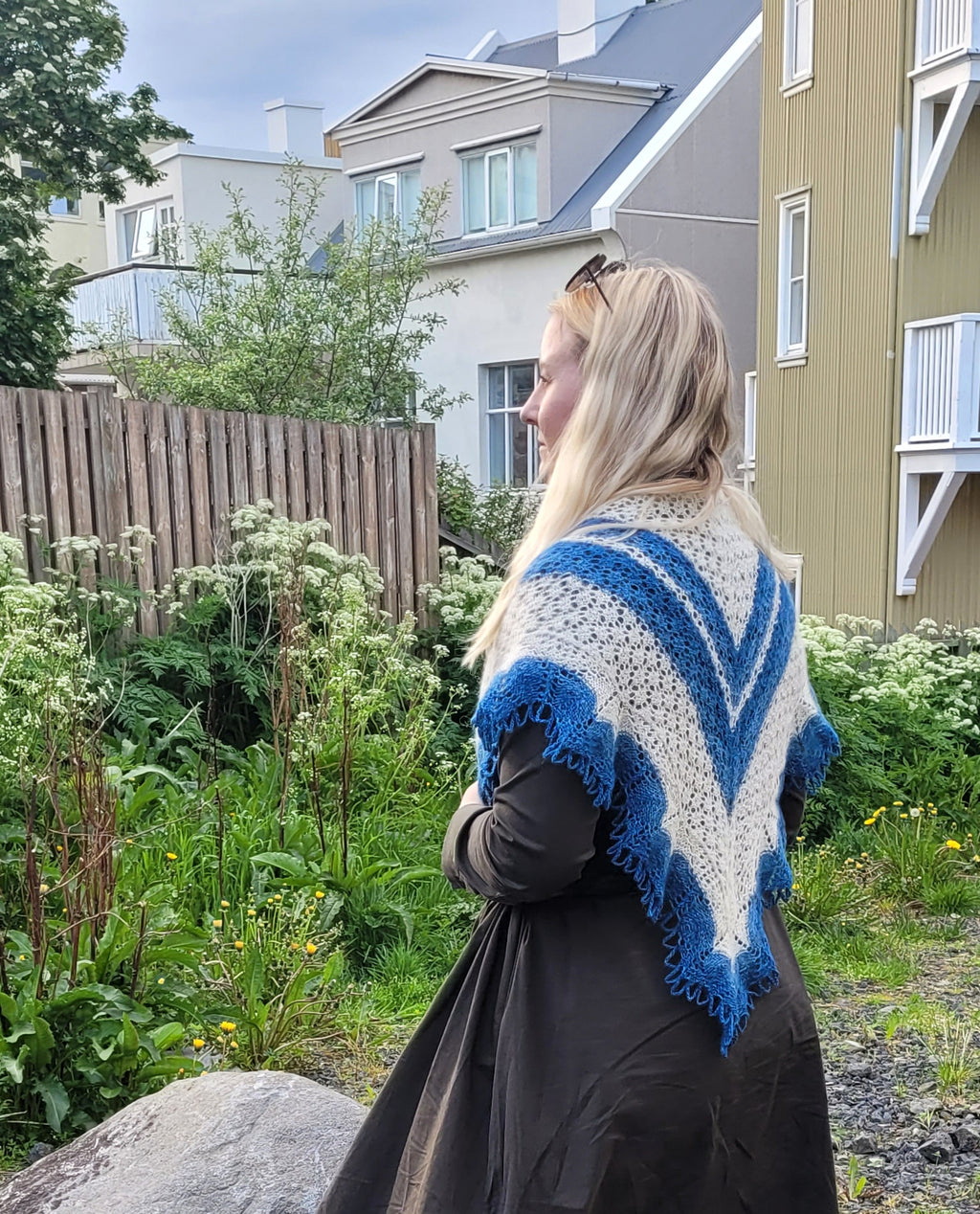Woman wearing a blue and white patterned shawl in an outdoor setting with houses and greenery.