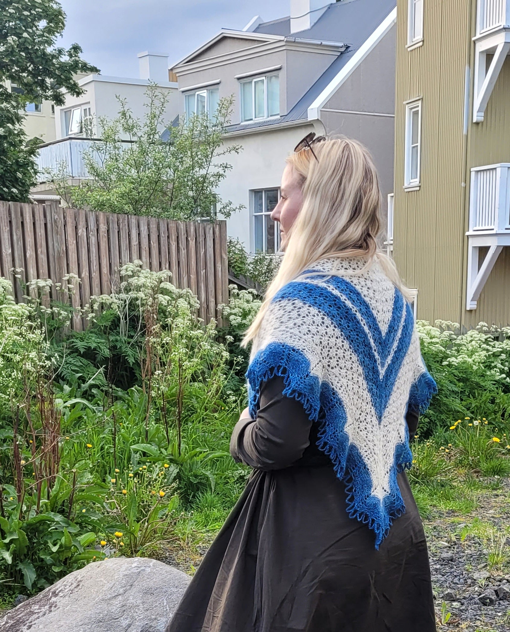 Woman wearing a blue and white patterned shawl in an outdoor setting with houses and greenery.
