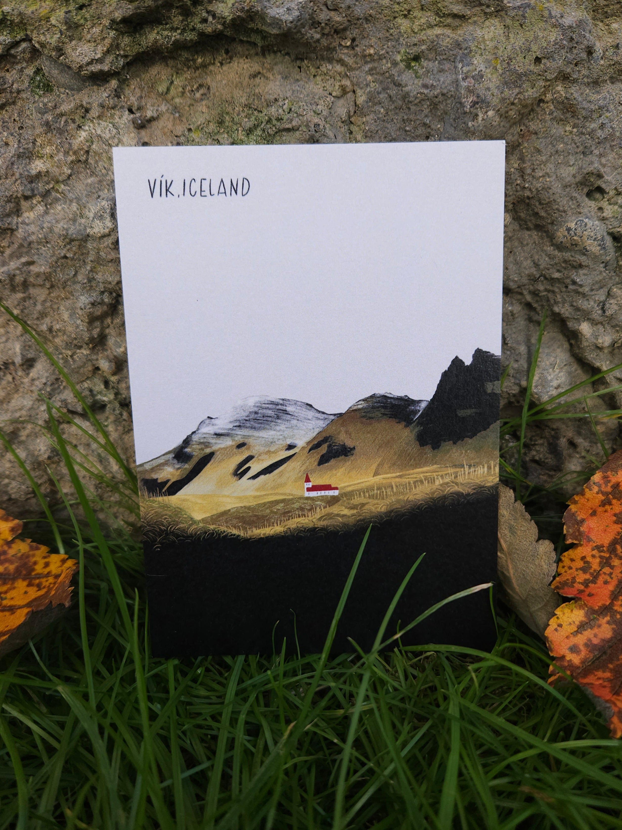 Postcard with mountain landscape design and 'Vik, Iceland' text on a stone surface with grass and leaves.