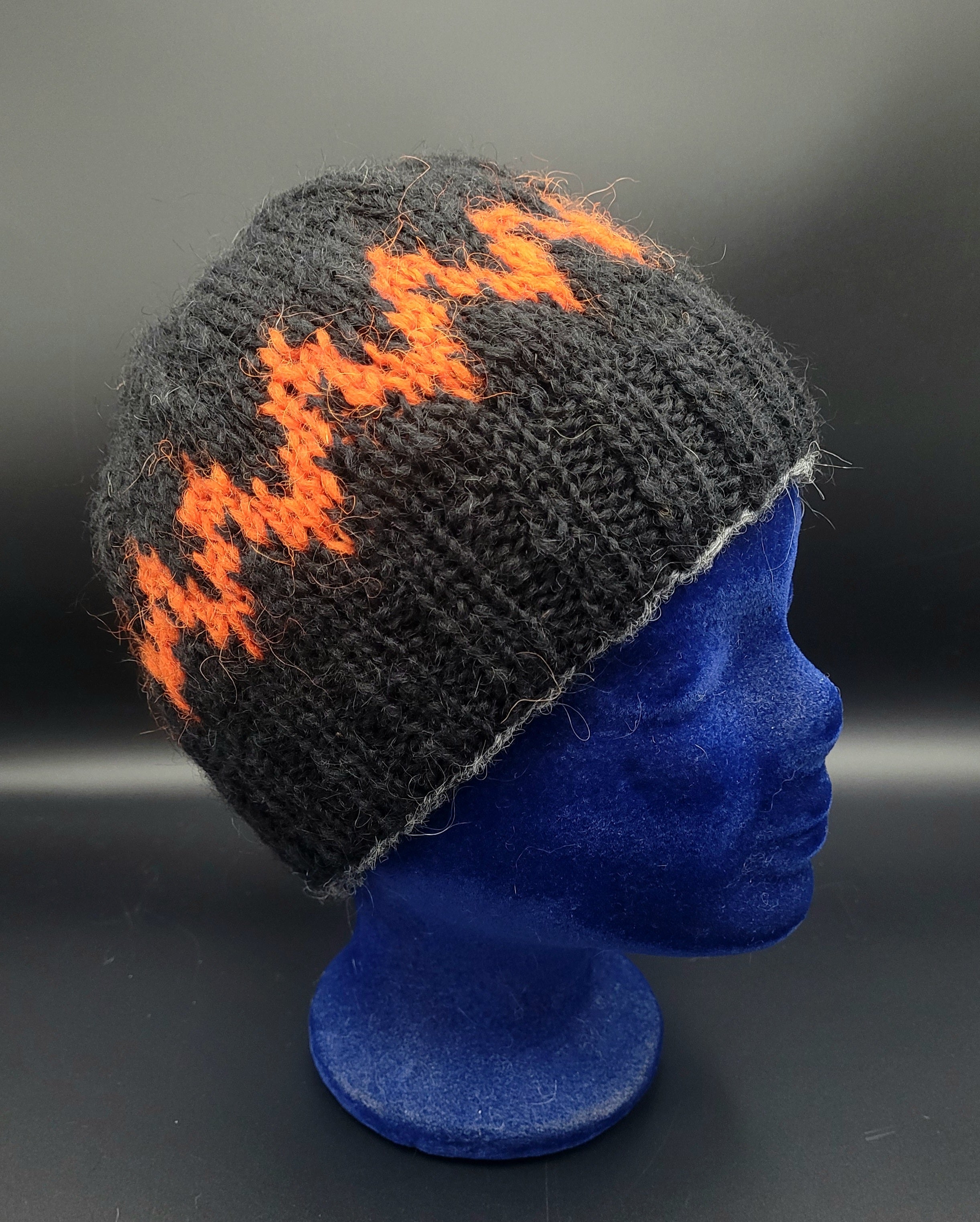 Black knit beanie with orange pattern on a blue mannequin head against a dark background