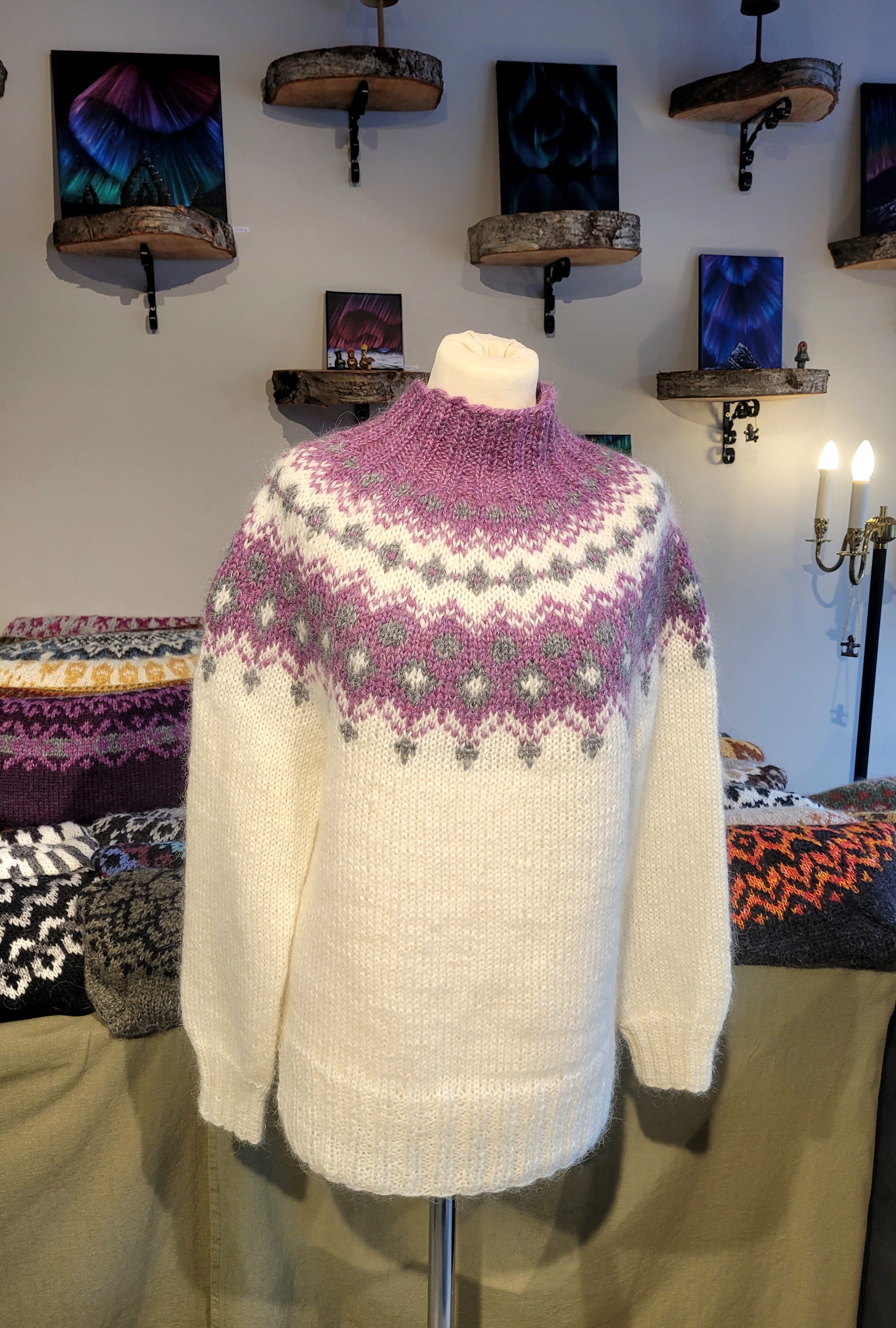White sweater with purple and green pattern on a mannequin against a wall with decorative items.